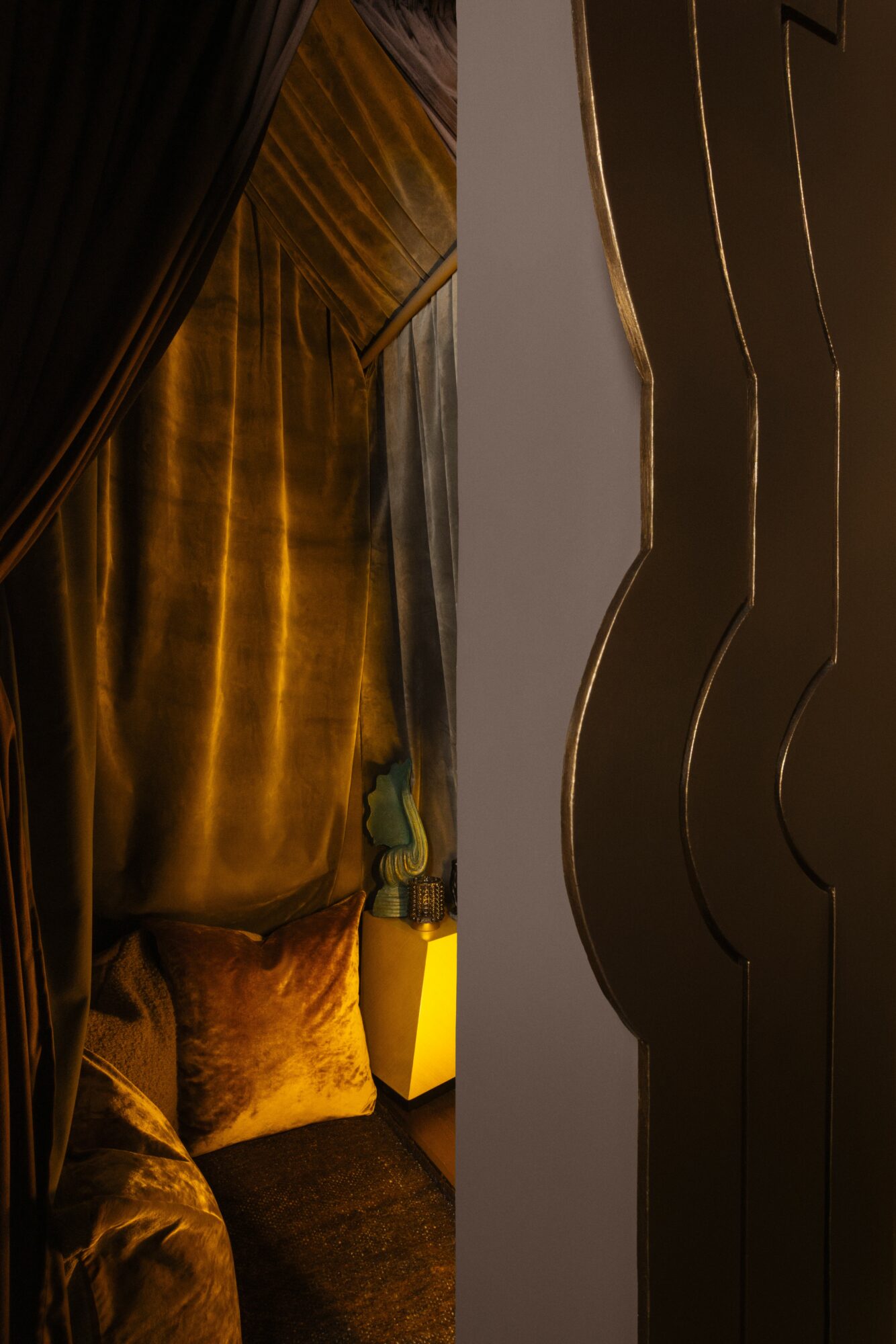 Part of a room with dark curtains, a golden wall, and a yellow lamp, viewed from the side.