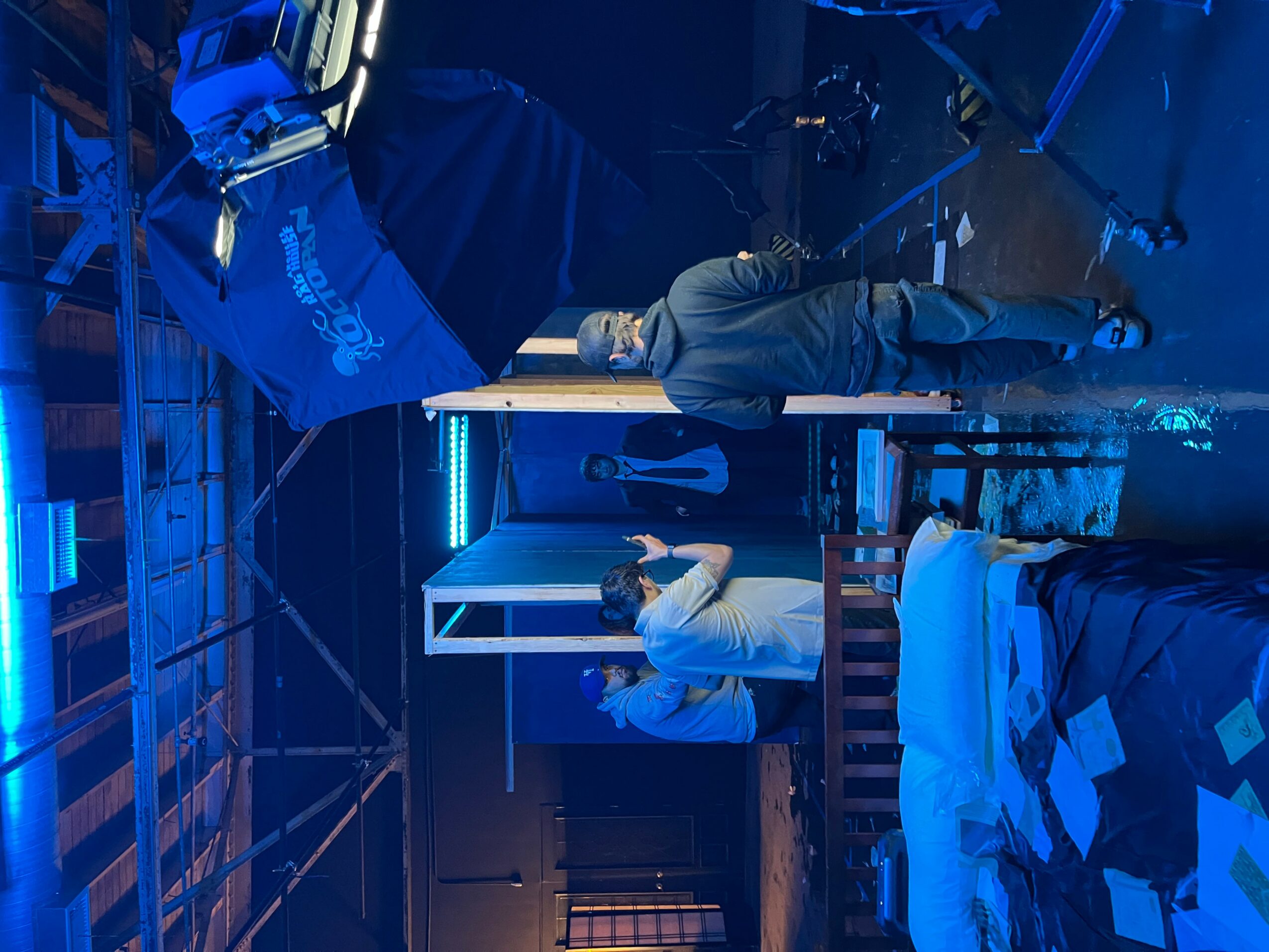 Two people working at a table in a room with blue lighting, one standing and one sitting, with a bed nearby.