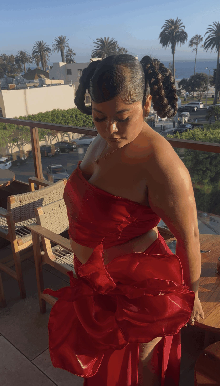 Young girl in a red dress holding a large red bow, standing outdoors with palm trees in background.