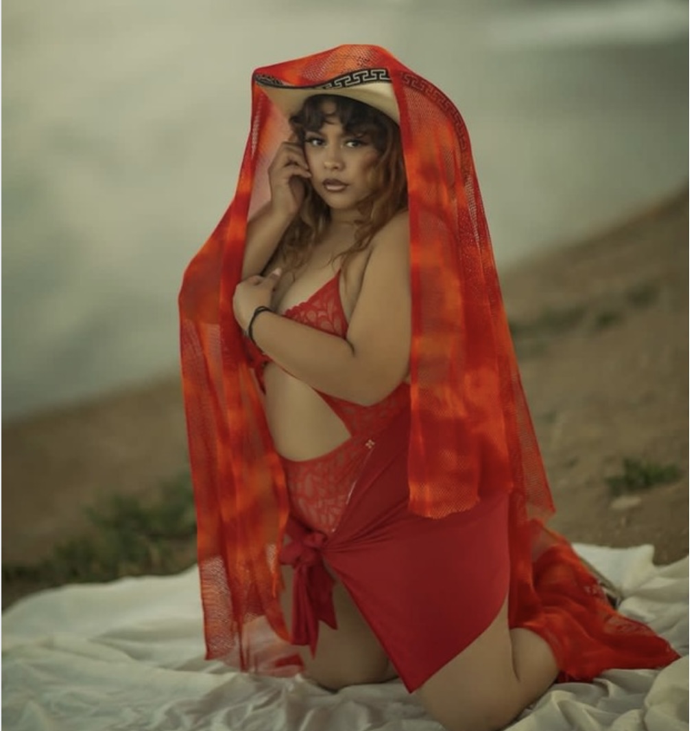 Woman in red clothing with a red sheer shawl, kneeling on a white cloth outdoors, holding her hair, with a blurred background.