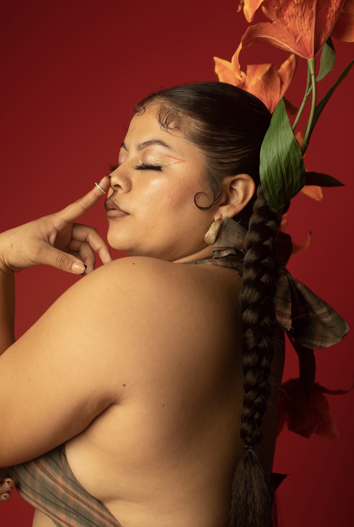 Woman with closed eyes and braided hair, holding finger to her nose, with red background and large orange flowers behind her.