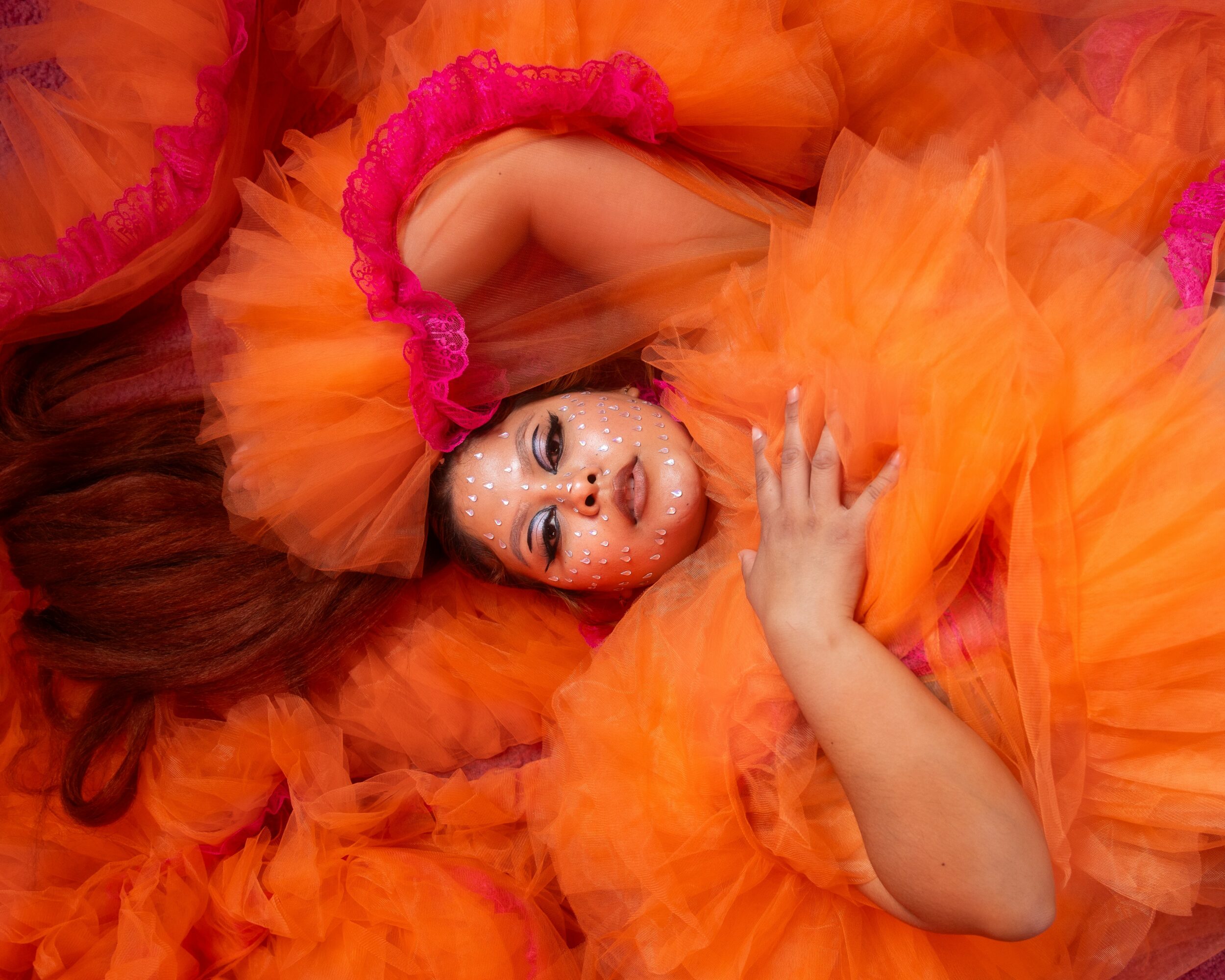 Person lying down in orange and pink ruffled costume, looking at camera, with hand near face.