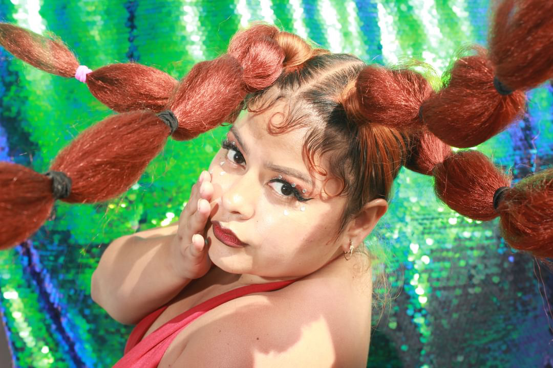 Child with large, puffy hair styled in multiple pigtails, touching face, against a colorful, shiny background.
