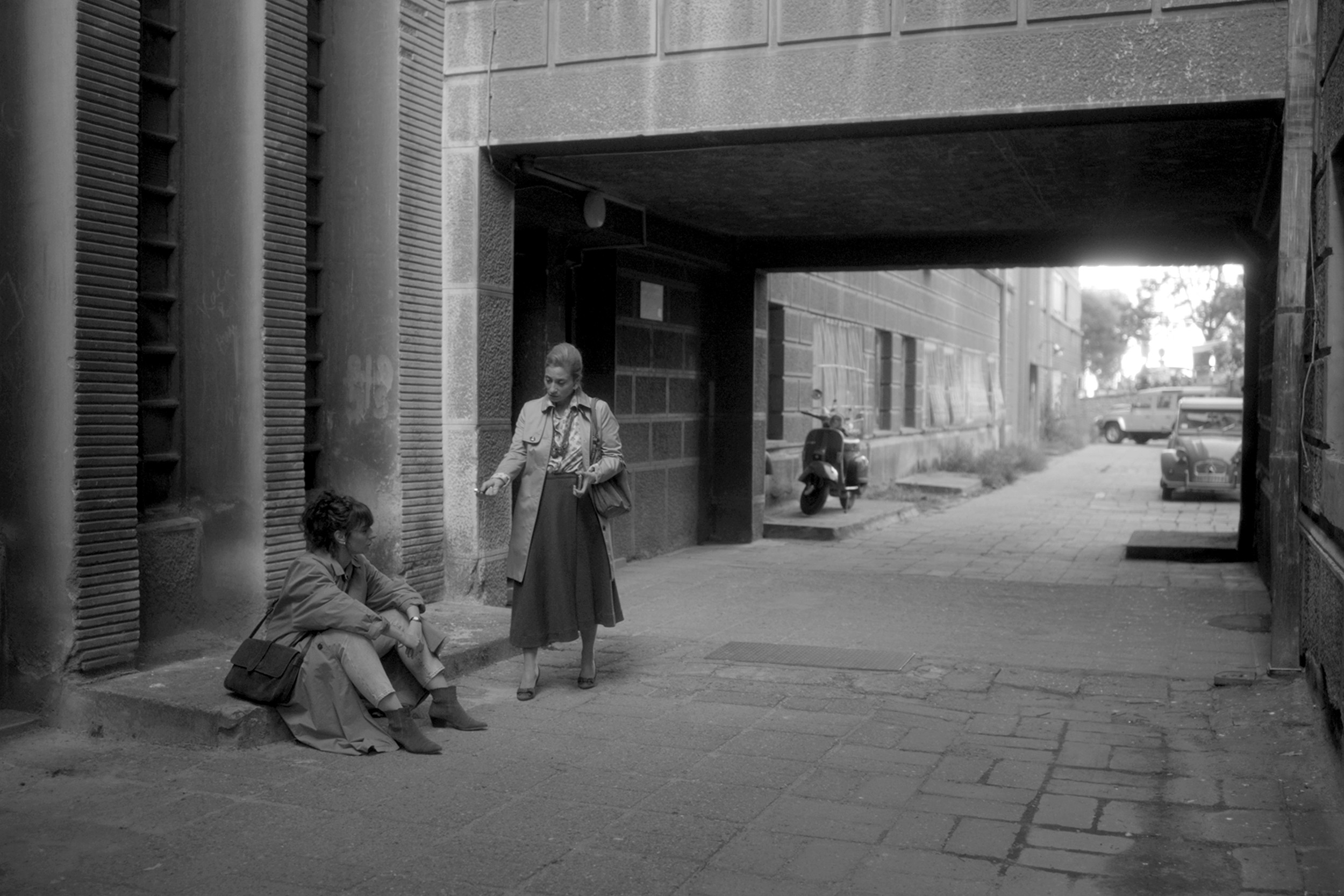 Two people sitting on the sidewalk and a woman standing nearby under an overpass in an urban setting.