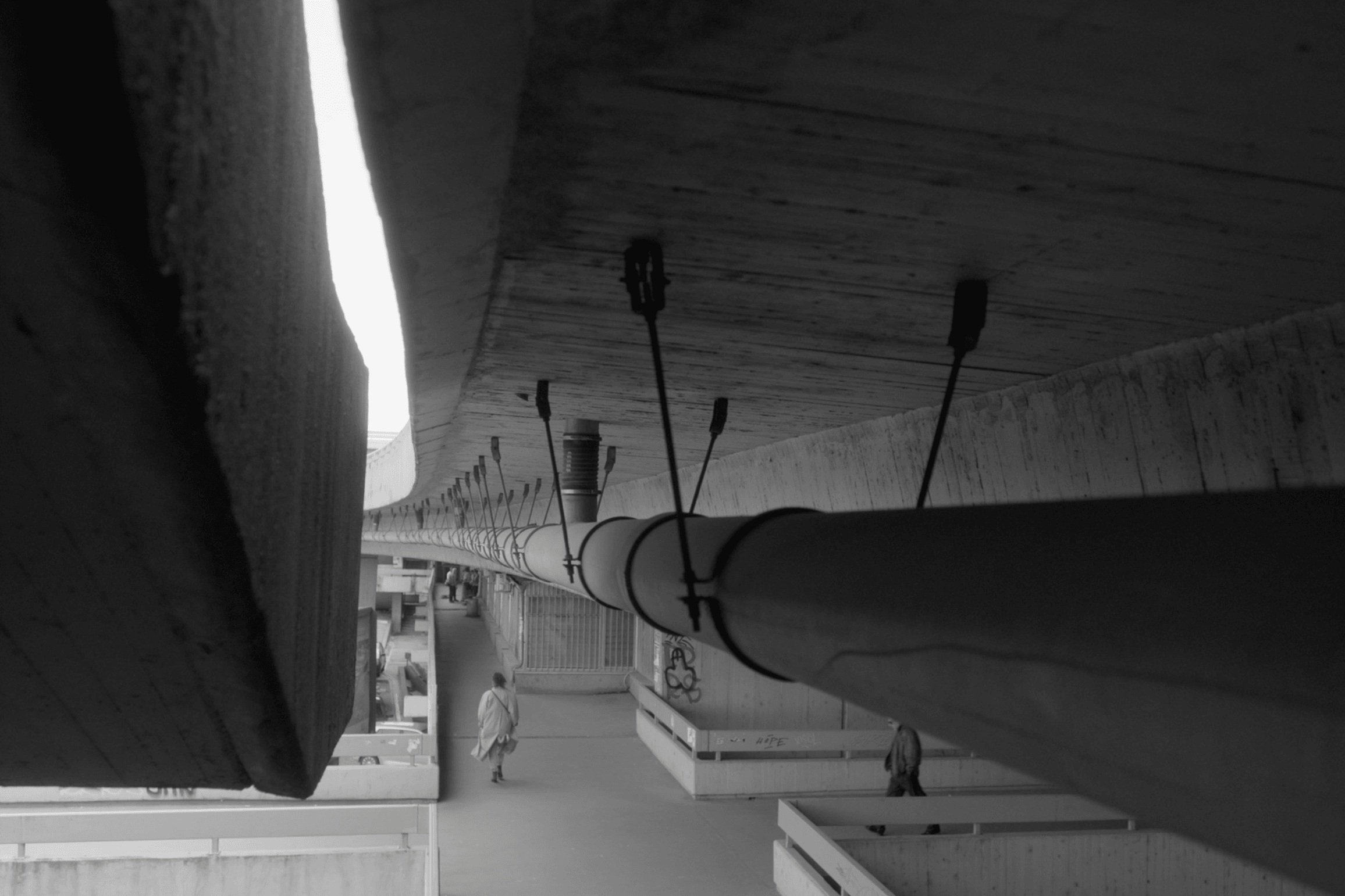 View of a large pipe with multiple smaller pipes and wires attached, seen from below in black and white.