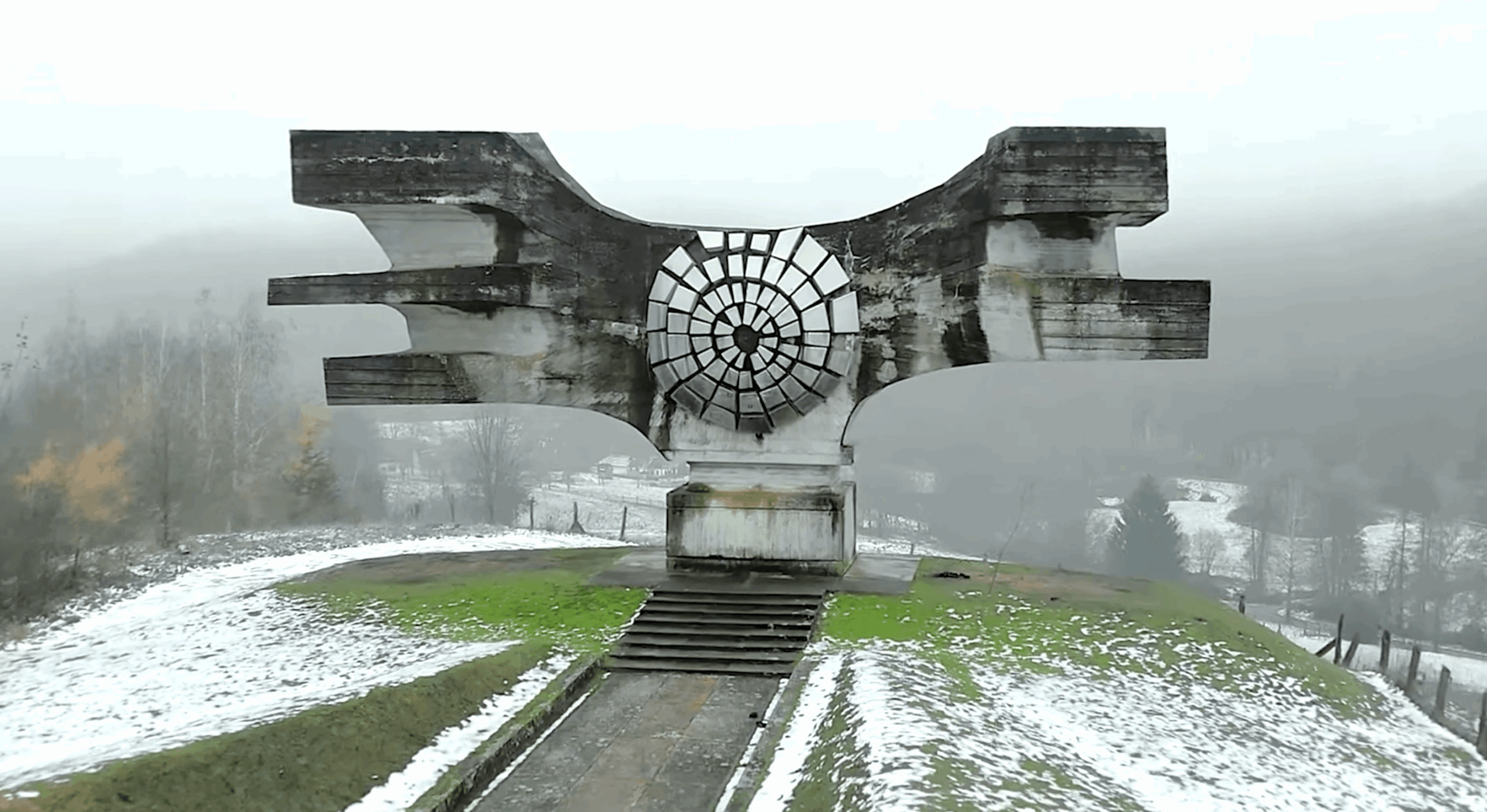 Concrete monument with a circular design, set on a grassy hill with snow, foggy background, and steps leading up to it.