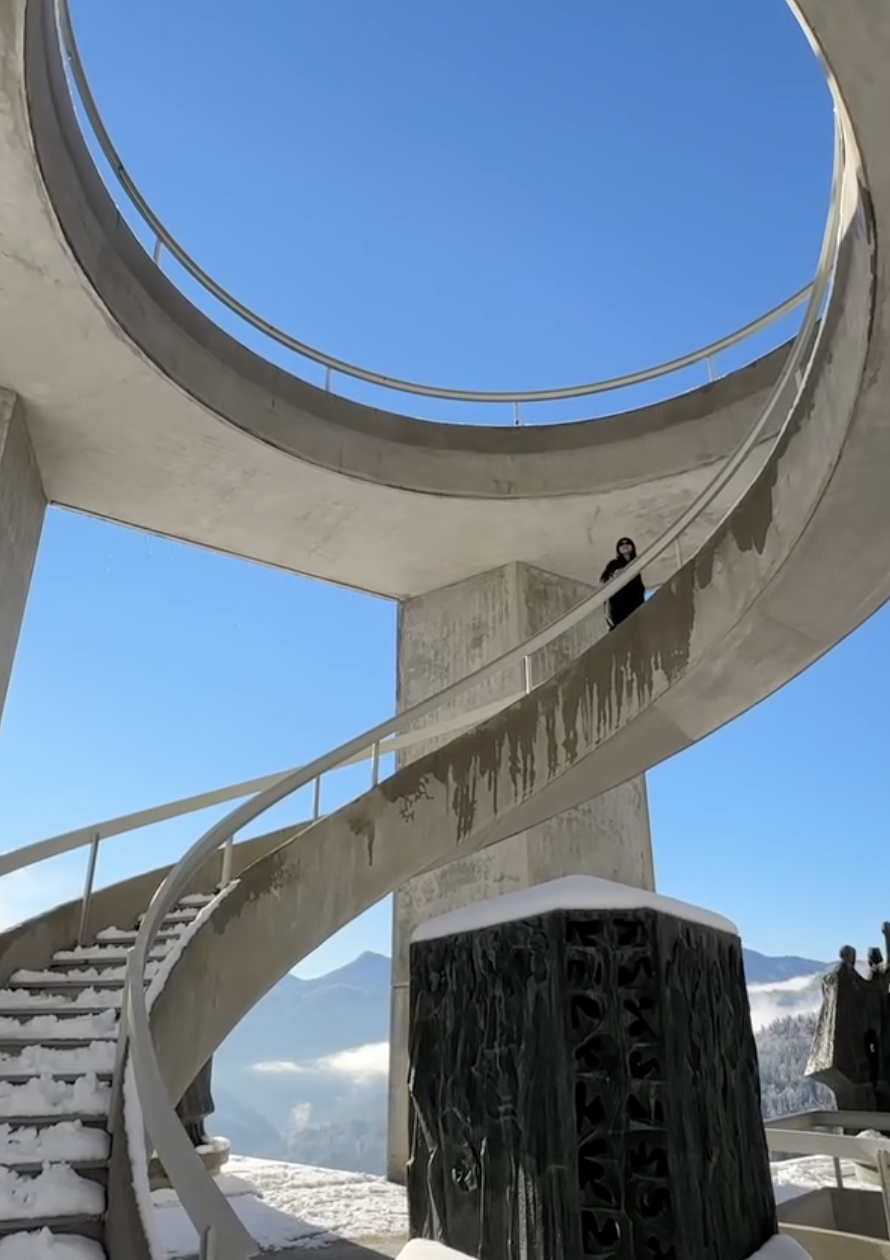 Spiral concrete staircase with a person standing on it, viewed from below against a blue sky with a circular opening.