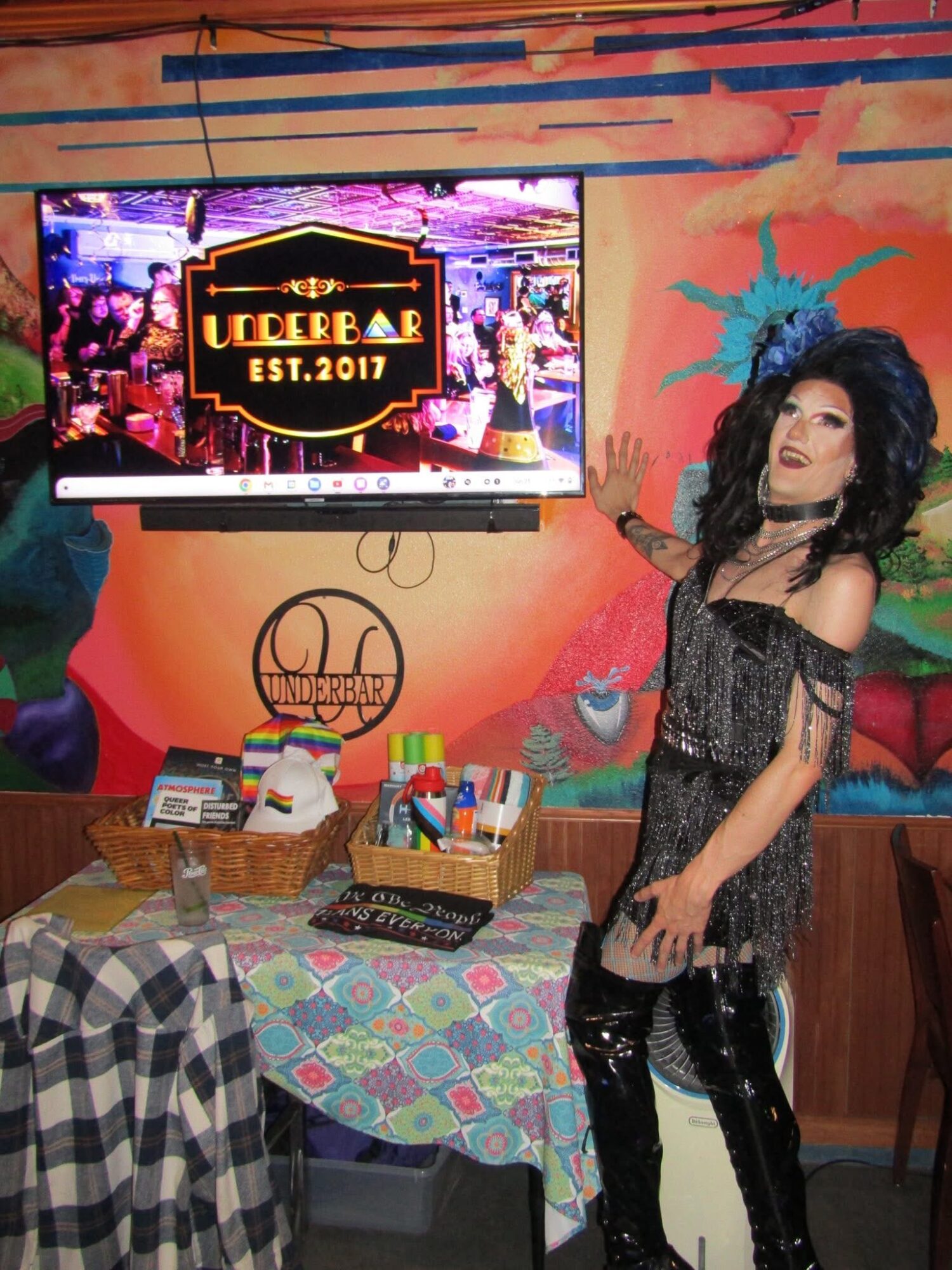 Person with curly hair in black outfit standing next to a table with art supplies, colorful wall background, and a screen displaying a logo.
