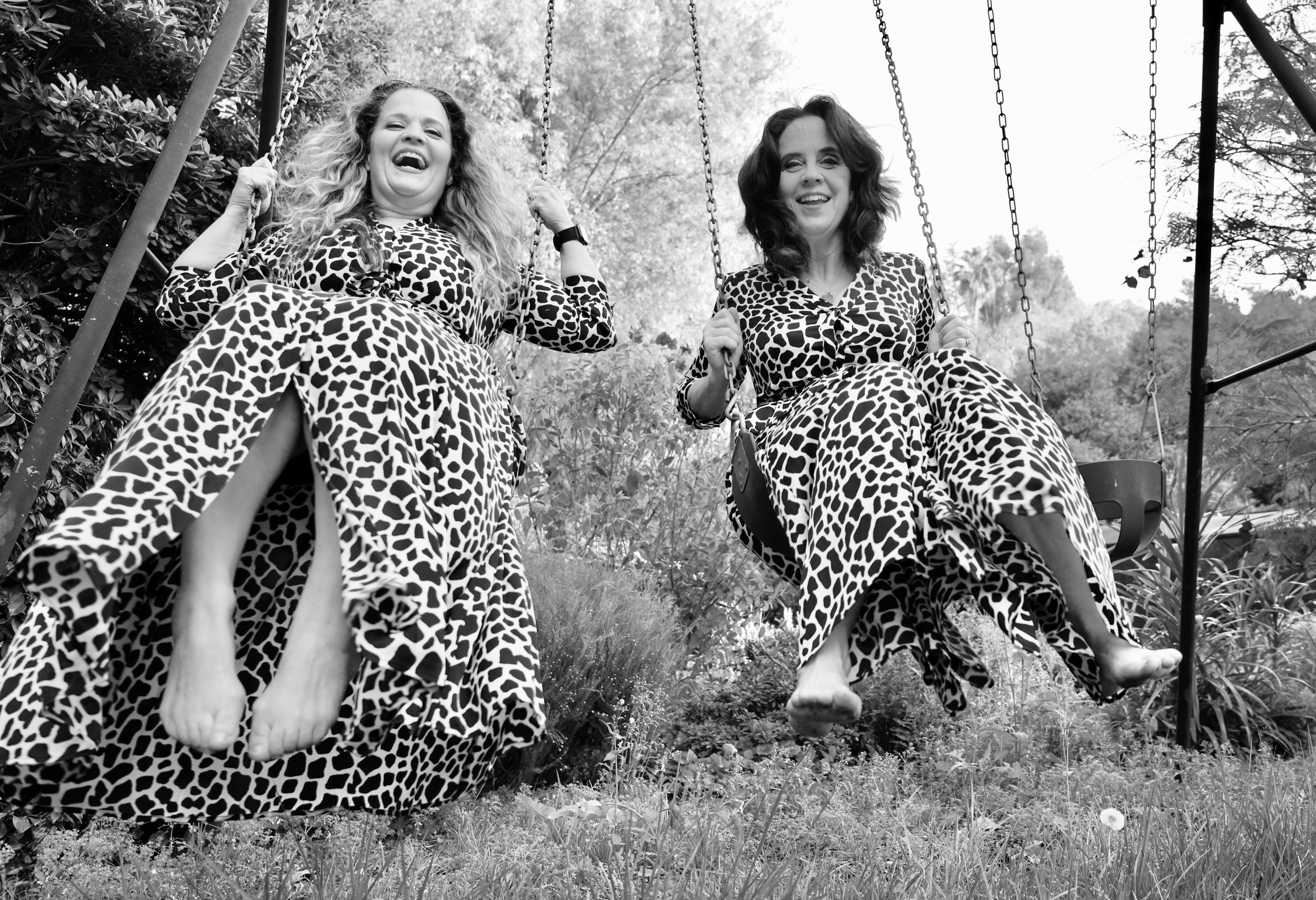 Two women on swings outdoors, smiling, wearing leopard print dresses, grass and trees in background.