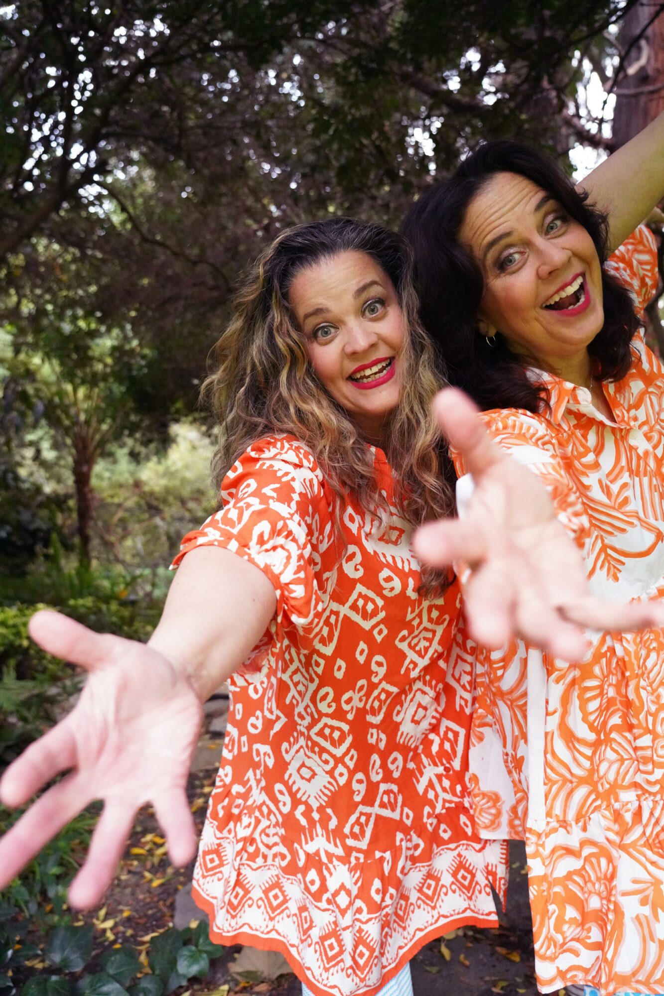 Two women in orange patterned dresses smiling and reaching toward the camera outdoors with trees in the background.