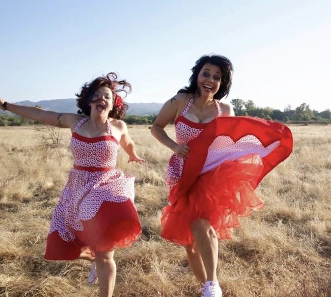 Two women running in a field, holding a red and white cloth, smiling, with mountains in the background.