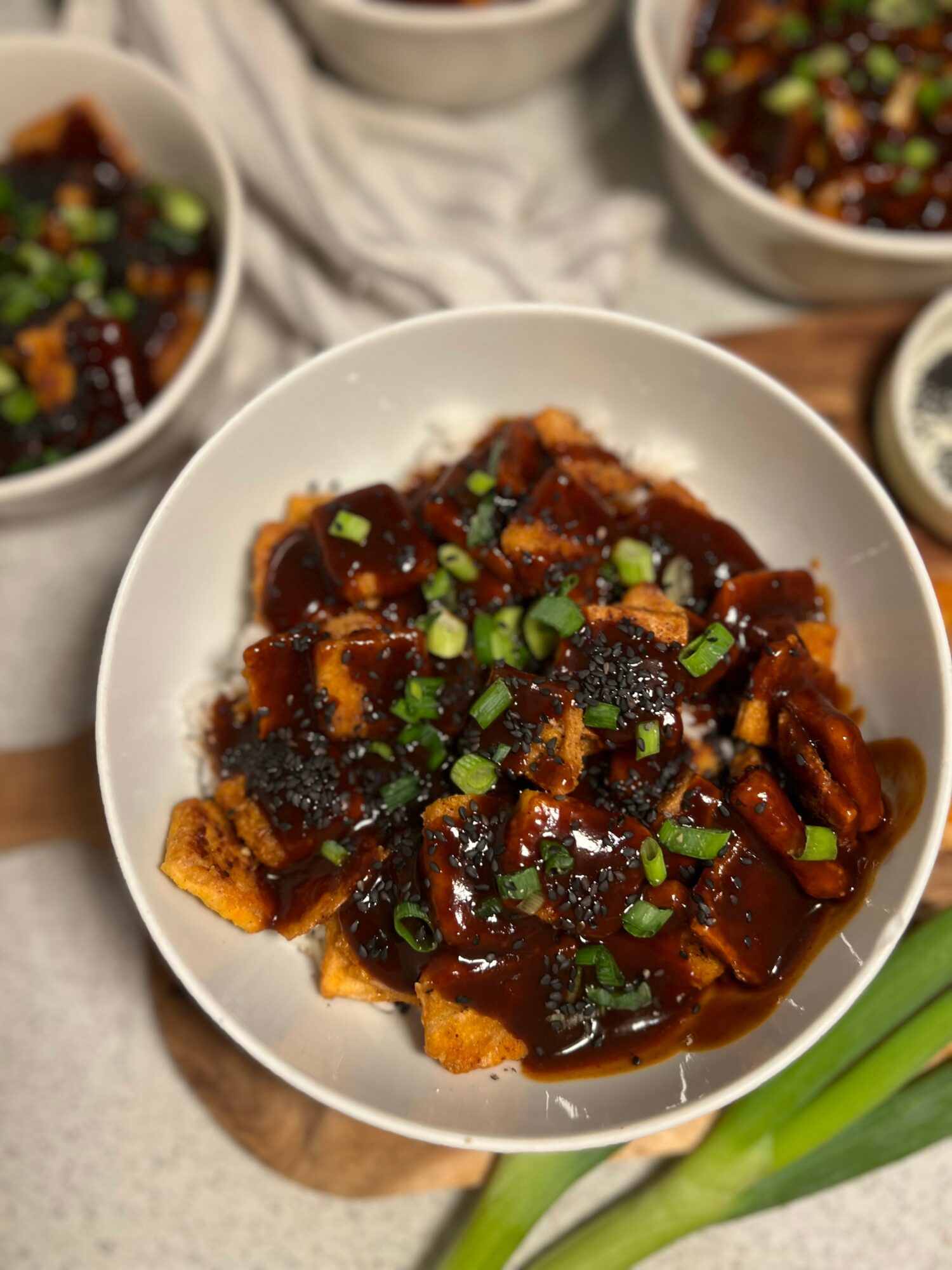 Bowl of tofu with dark sauce and chopped green onions, surrounded by other bowls of similar dishes, on a wooden surface.