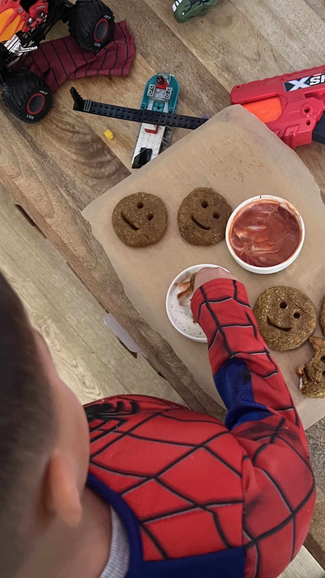 Child in Spider-Man costume reaching for cookies and ketchup on a wooden surface, with toys and tools nearby.