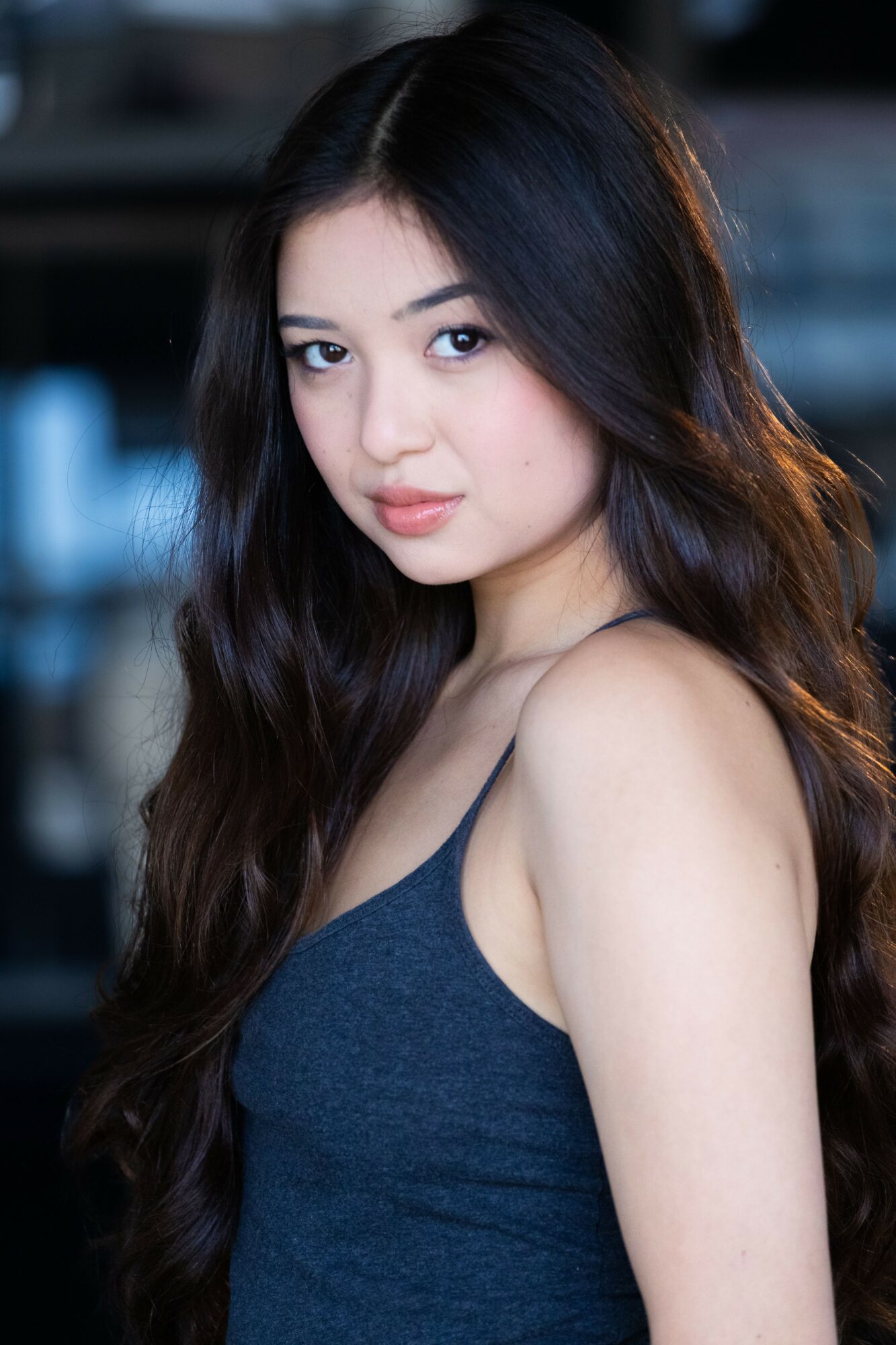 Young woman with long dark hair and a dark tank top, looking at the camera, in an indoor setting.