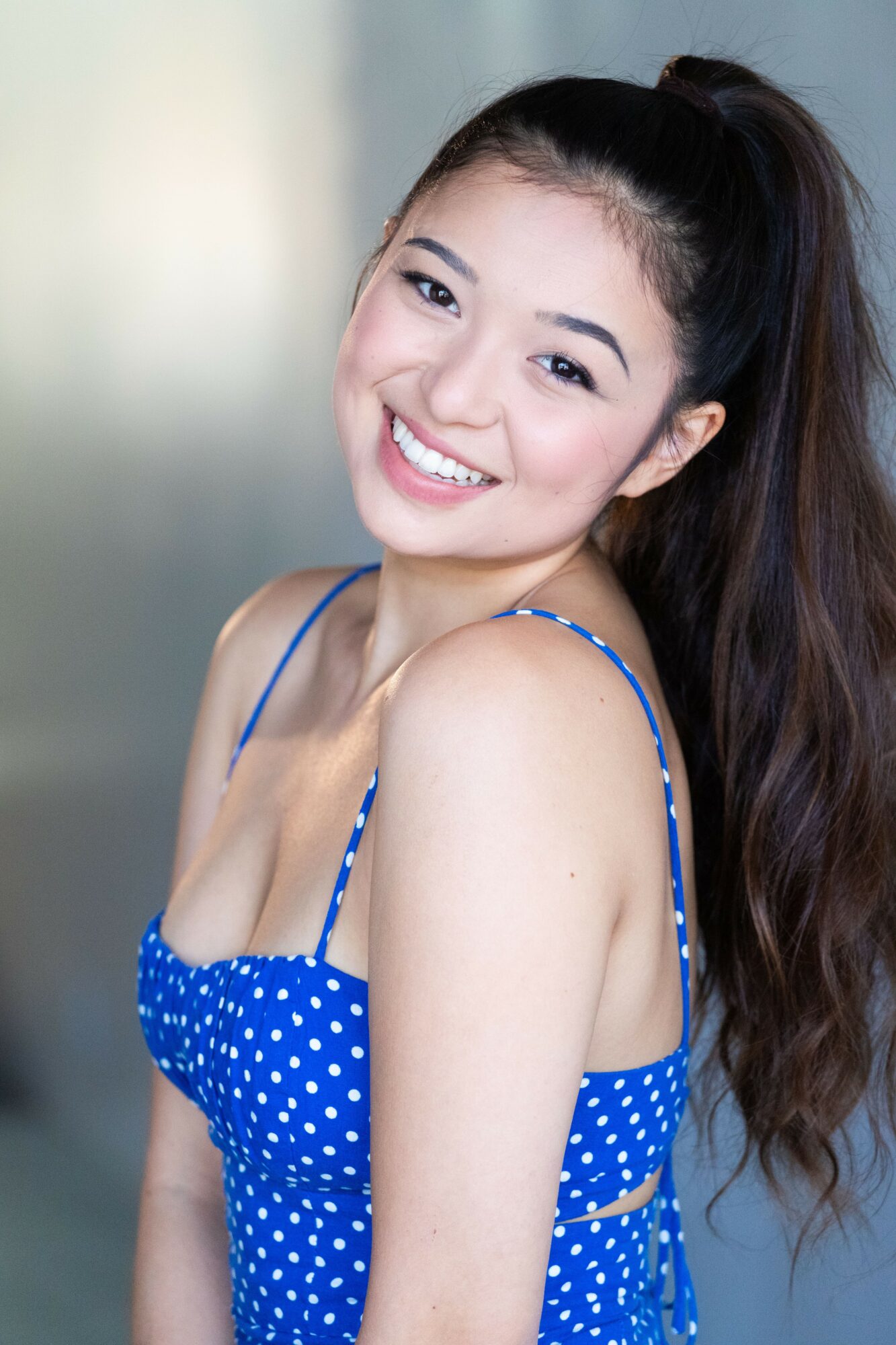 Smiling young woman with long dark hair in a blue polka dot dress, looking at camera.
