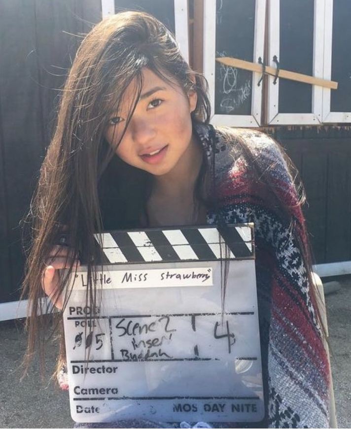 Young woman with long hair holding a clapperboard outdoors, sunlight on her face, dark background, wearing a patterned sweater.