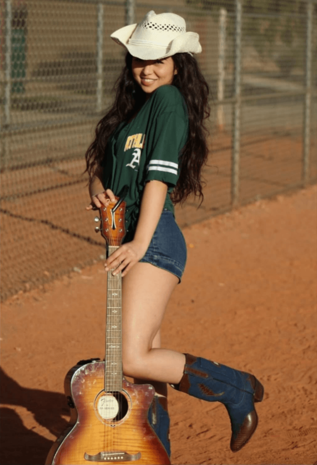 Young woman in a green sports jersey, denim shorts, cowboy hat, and boots, holding a guitar on a tennis court.