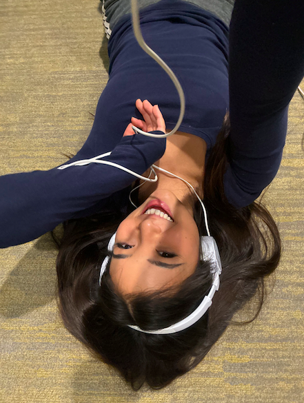 Young woman lying on the floor, smiling, wearing headphones, with a hand near her face, on a beige carpet.