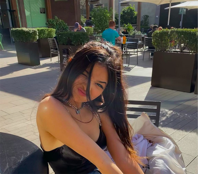 Young woman with long dark hair smiling outdoors, sitting at a table with potted plants and people in the background.