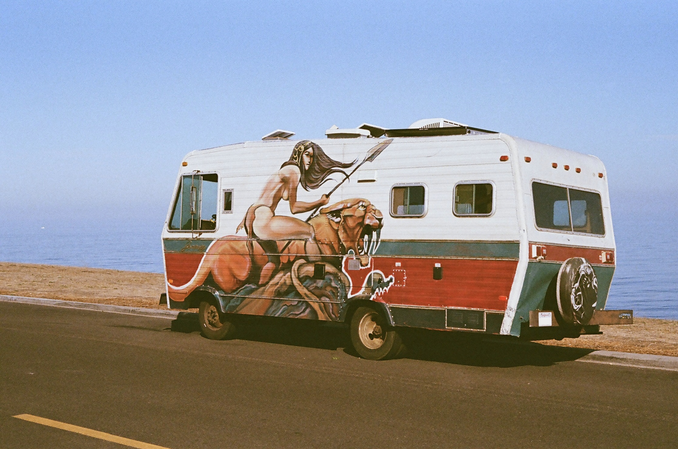 Camper van with a mural of a woman riding a tiger painted on its side, parked near a beach under a clear sky.