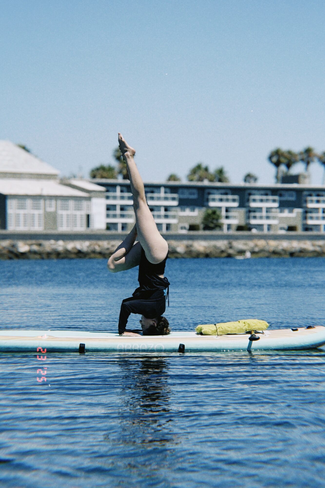 Person performing a headstand on a paddleboard in water, with buildings and trees in background.