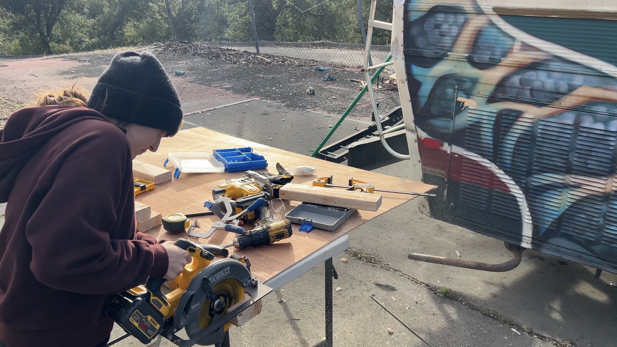 Person wearing a black beanie and maroon hoodie working at a table outdoors with tools and wood pieces.
