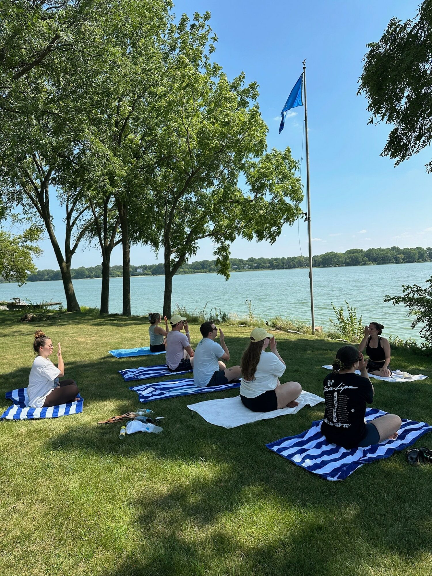 People sitting on striped blankets near a lake, trees, and a flagpole with a blue flag, under a clear sky.