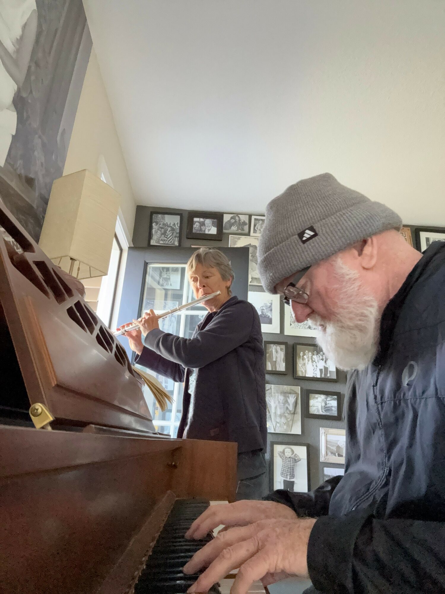 Two elderly men playing piano in a room with framed photos on the wall, one wearing a gray beanie and glasses.