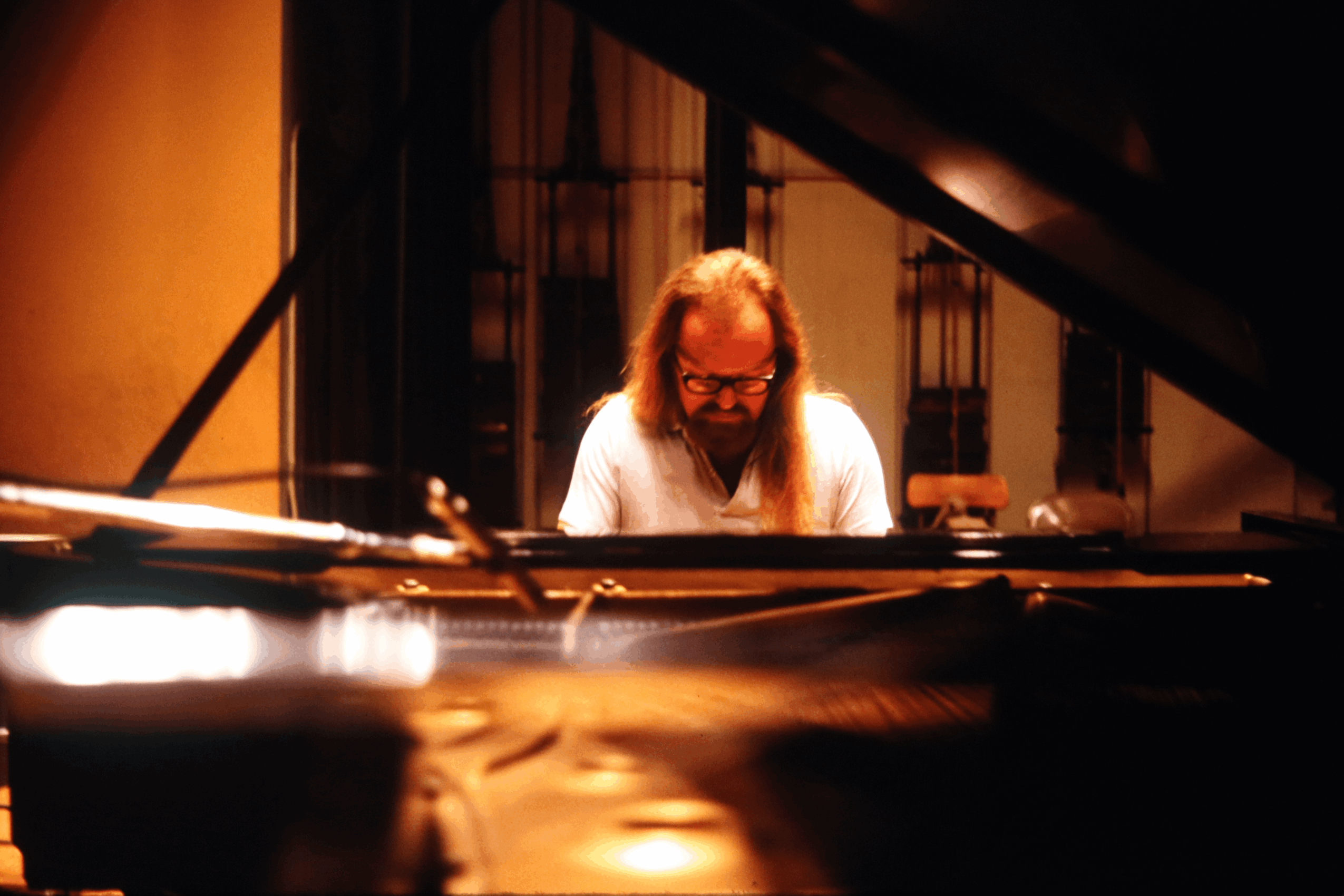 Person with long hair and glasses playing a grand piano in a dimly lit room.