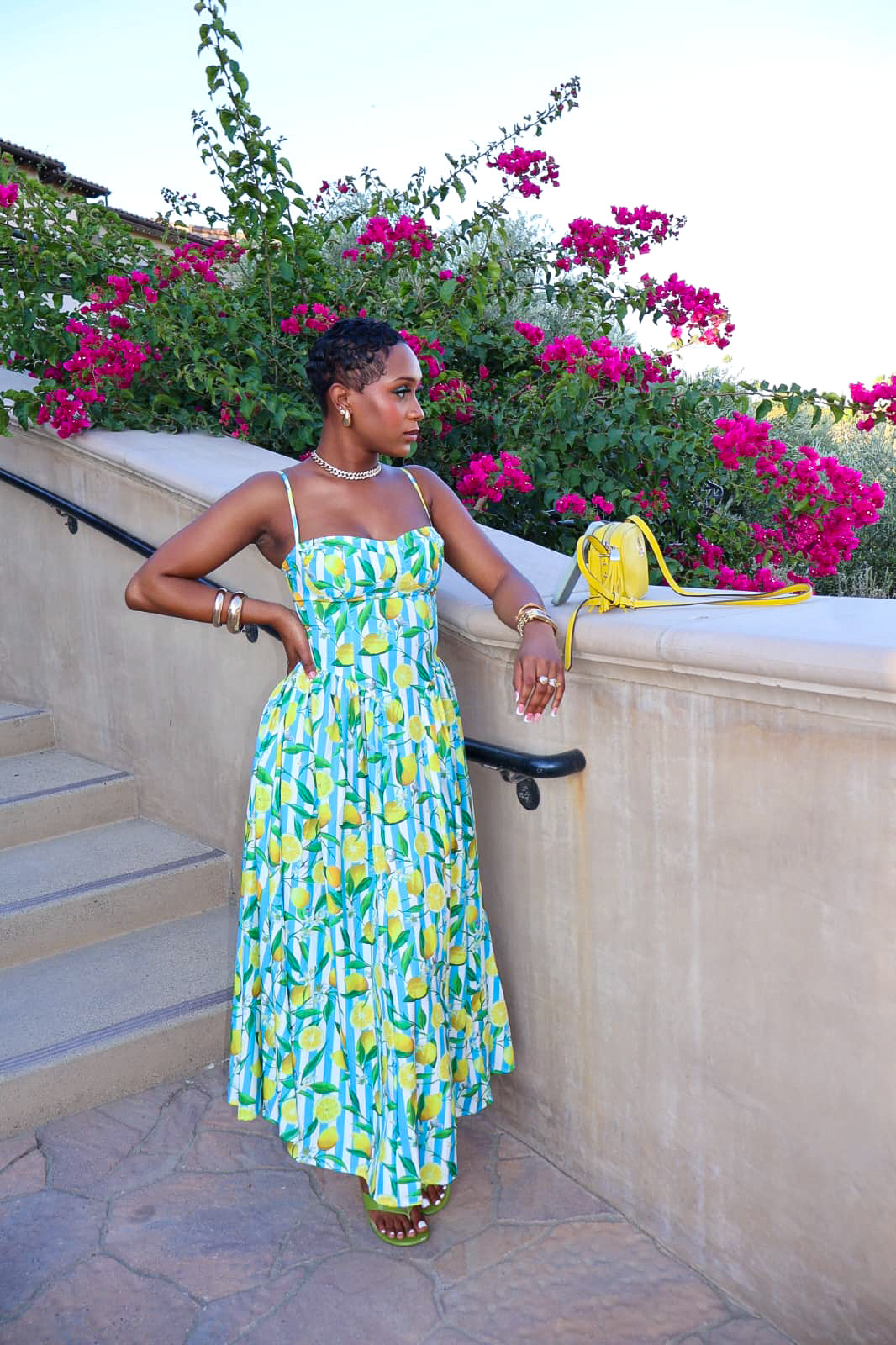 Woman in a colorful dress with a yellow bag, standing near a stone railing with pink flowers in background.