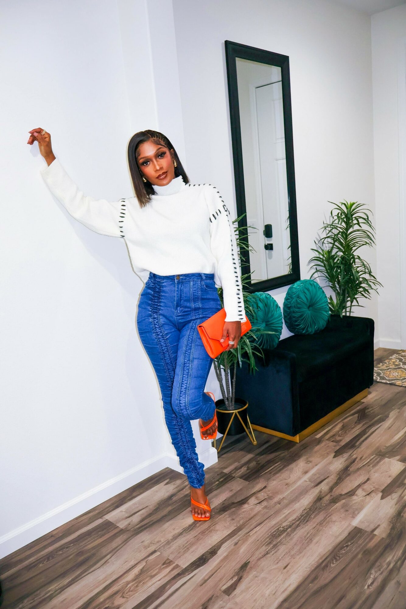 Woman in white sweater and blue jeans standing indoors, holding an orange clutch, with a mirror and plant behind her.