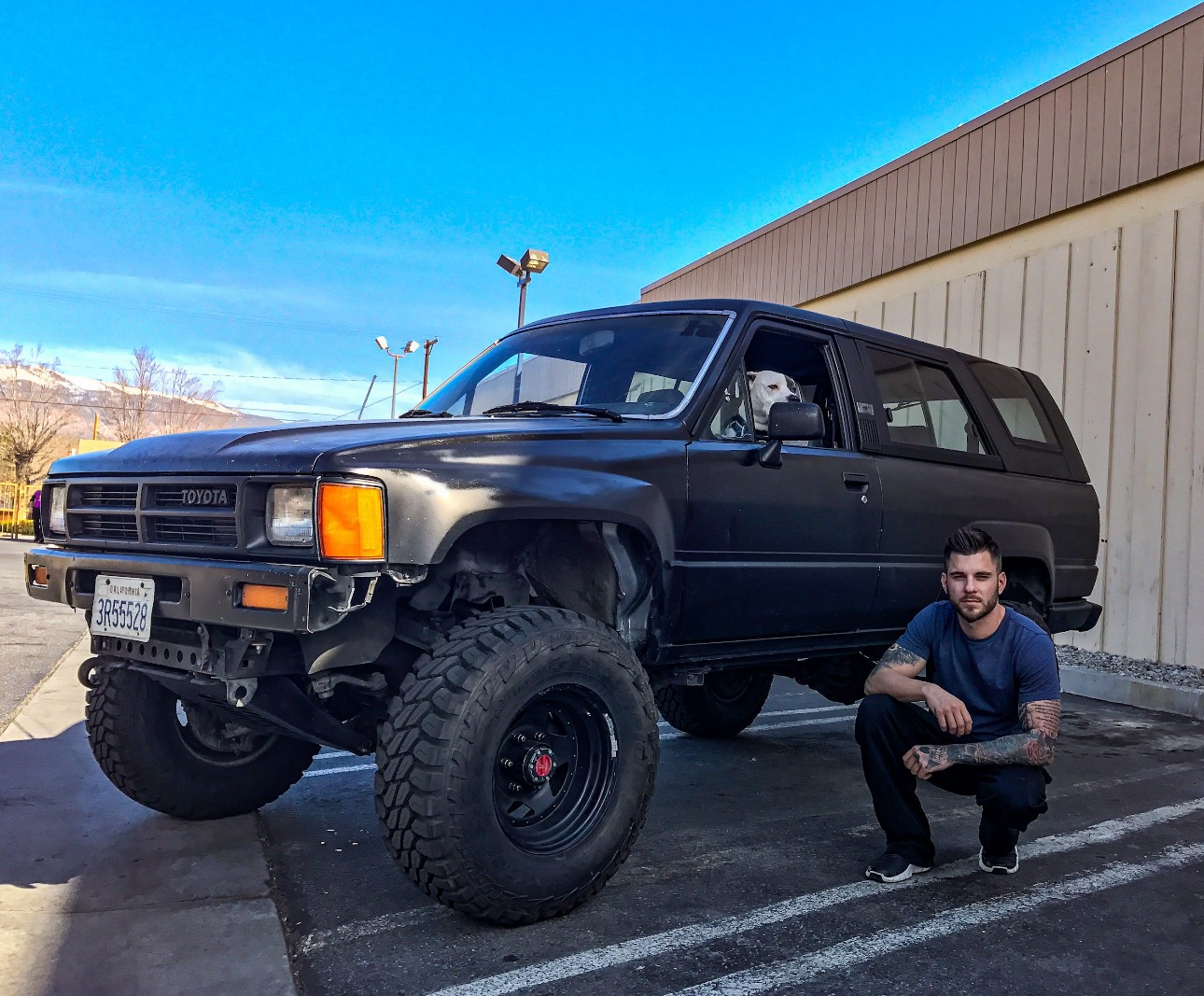 Person crouching next to a large black off-road vehicle parked outside a building under a blue sky.