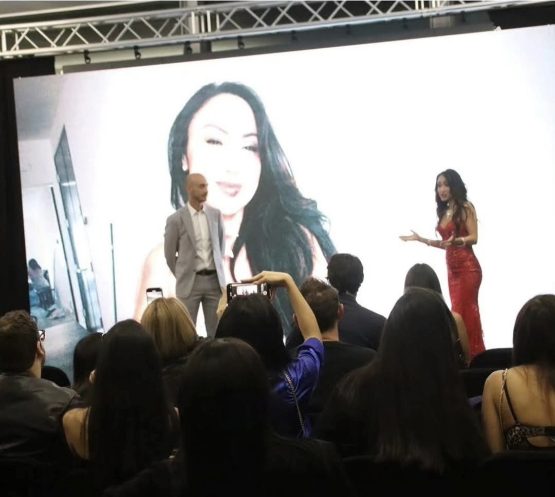 Two people on stage in front of a large screen with a woman's face, audience watching, one person taking a photo.