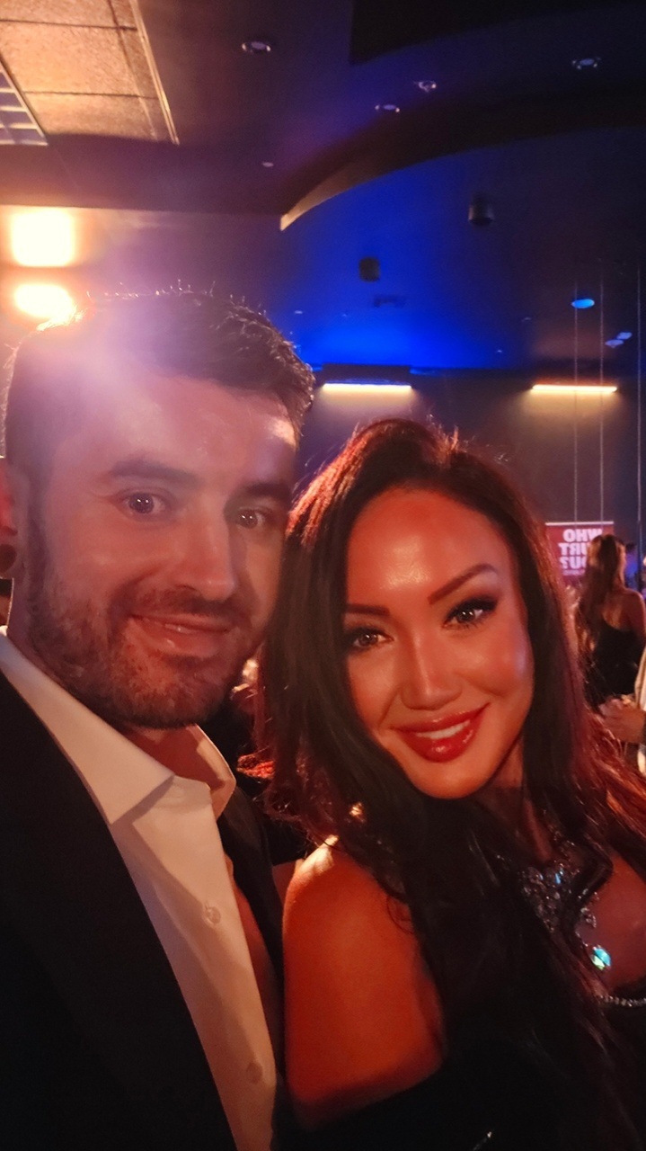 Two people smiling at a social event with colorful lighting and a blue ceiling in the background.