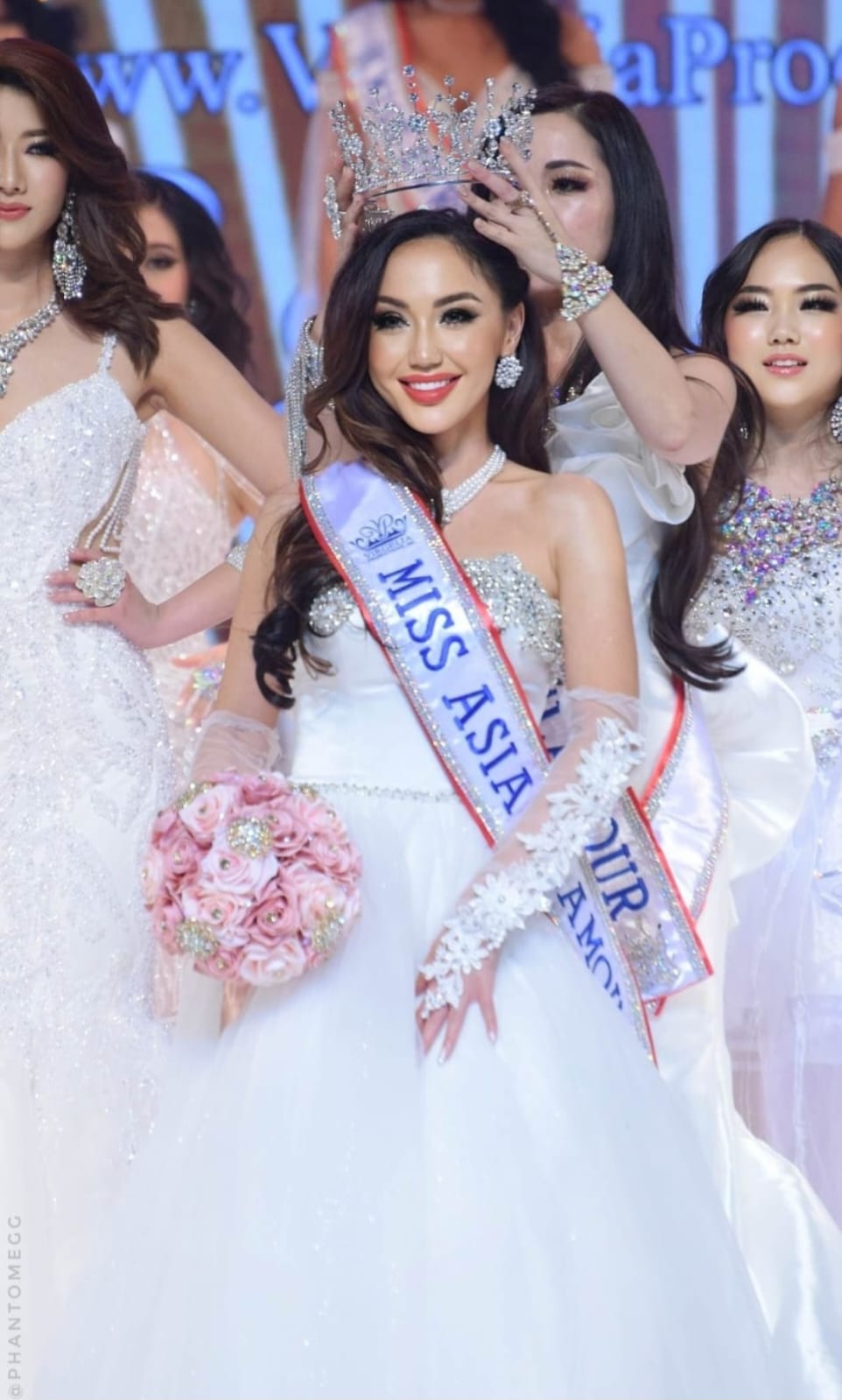 Beauty pageant winner in a white gown with sash, holding pink flowers, smiling on stage with other contestants behind her.