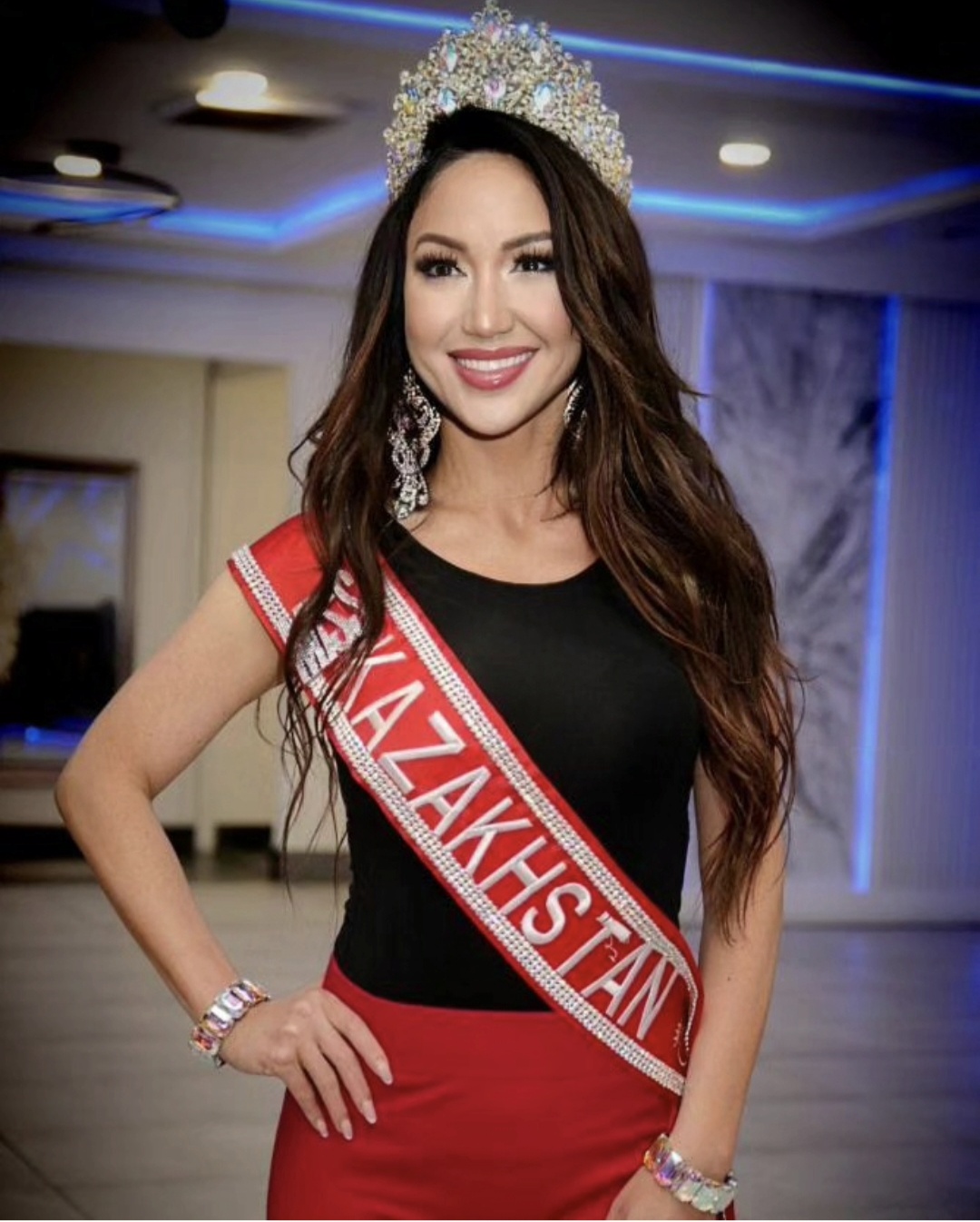 Woman wearing a crown and sash, smiling, in an indoor setting with blue lighting, long dark hair, and jewelry.
