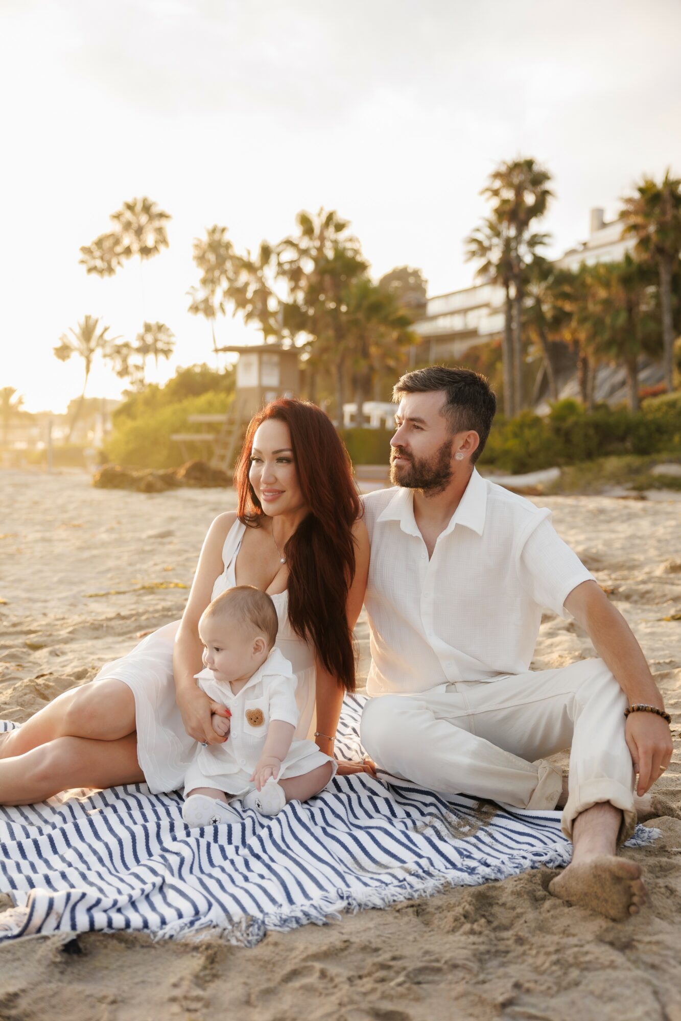 Family sitting on a striped blanket on the beach with palm trees and sunset in background, woman holding a baby, man beside her
