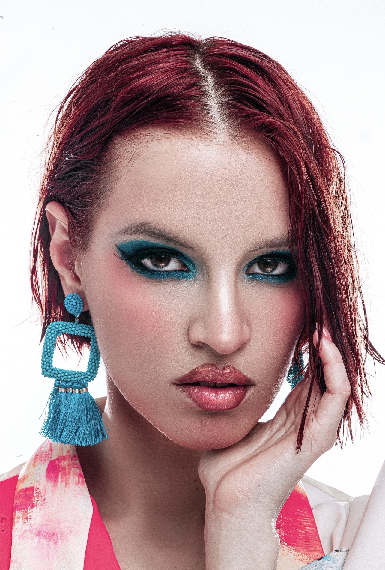 Close-up of a woman with red hair, bold makeup, and large turquoise earrings, touching her face gently.