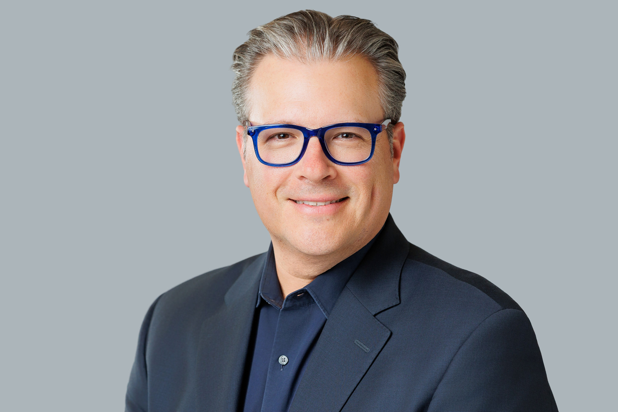 Man with gray hair, glasses, wearing a dark suit and shirt, smiling against a light gray background.