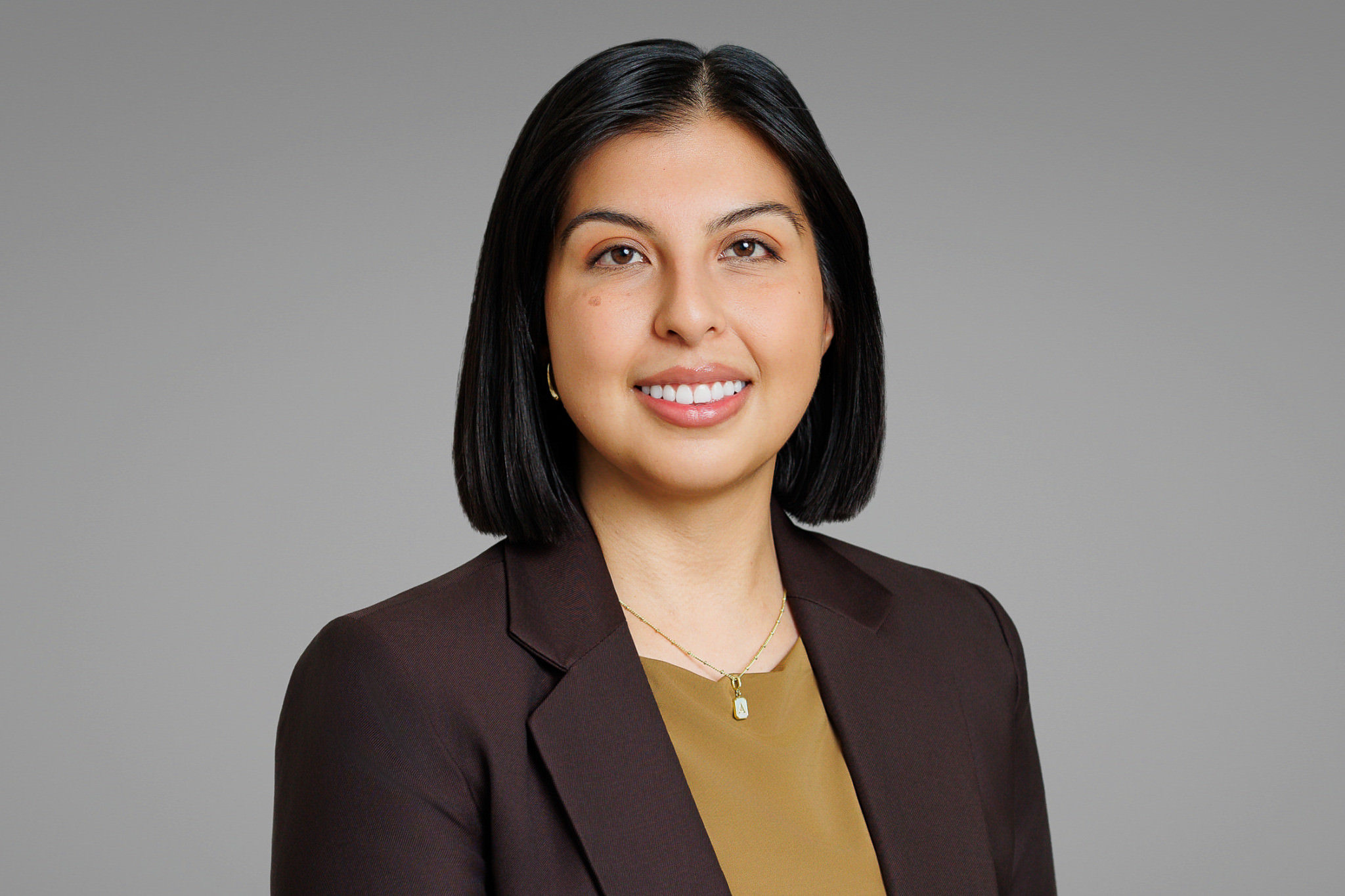Young woman with shoulder-length black hair smiling, wearing a dark blazer and a gold top, against a gray background.
