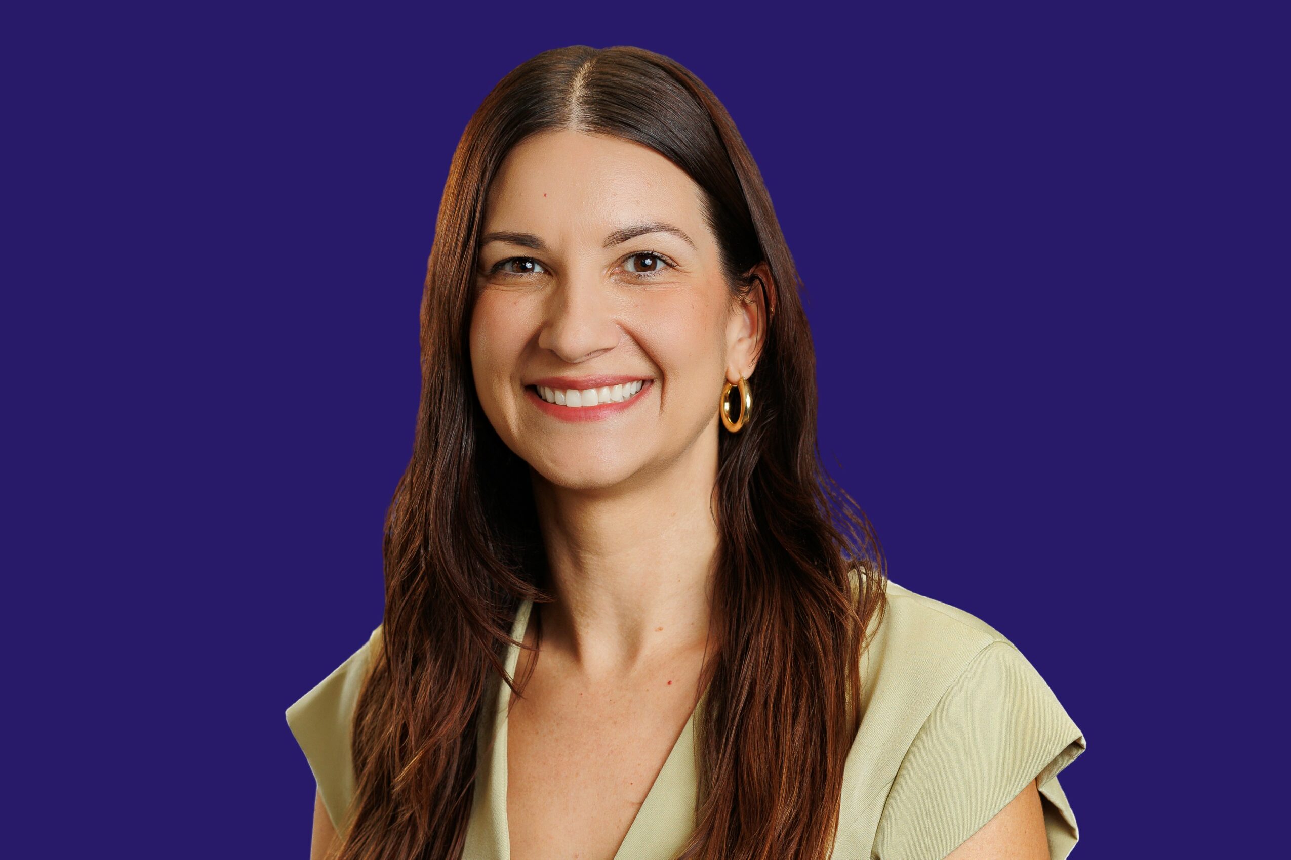 Smiling woman with long brown hair, wearing a light-colored top and gold hoop earrings, against a dark blue background.