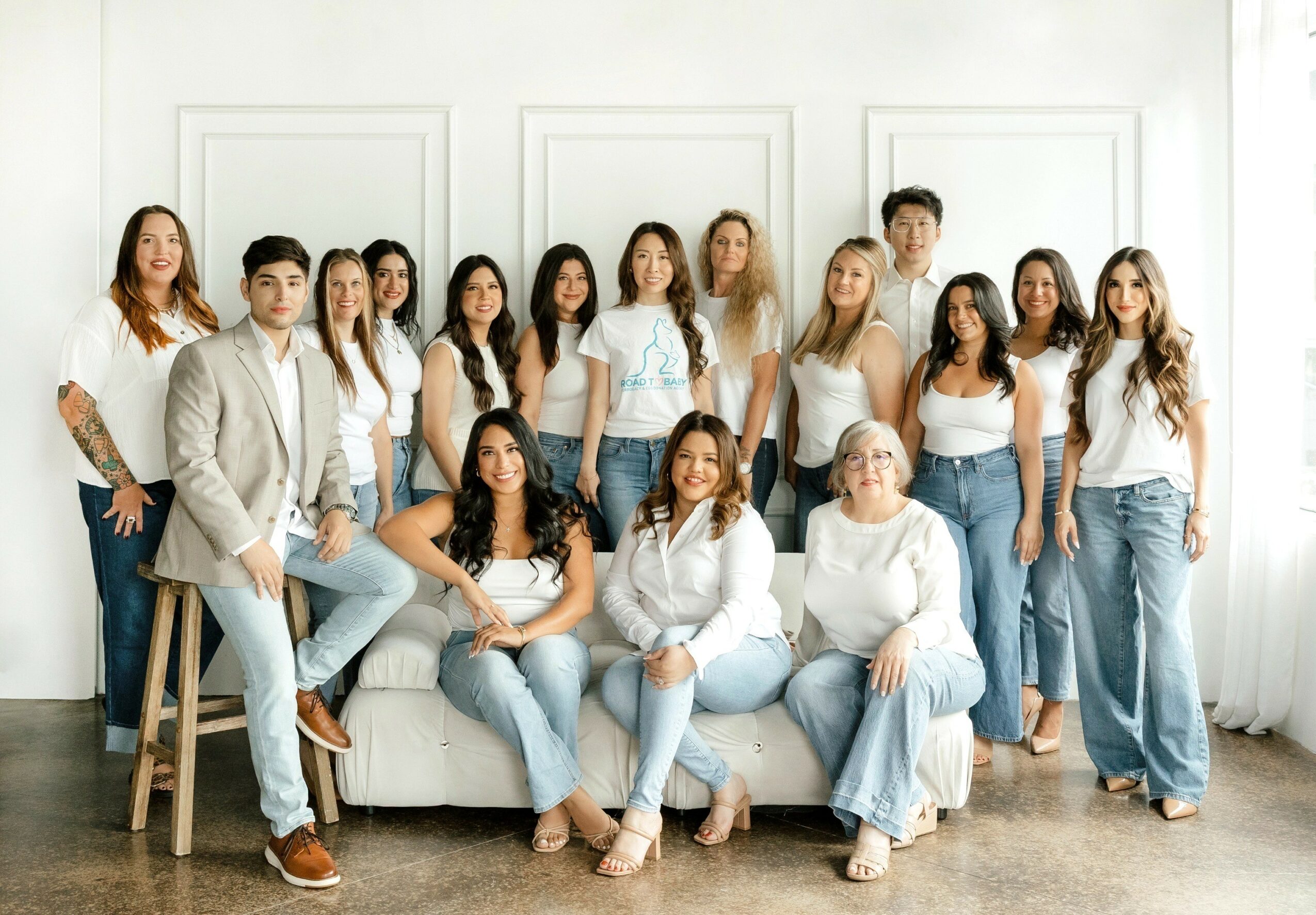 Group of 16 people, mostly women, posing in a bright room with white walls and curtains, some sitting and some standing.