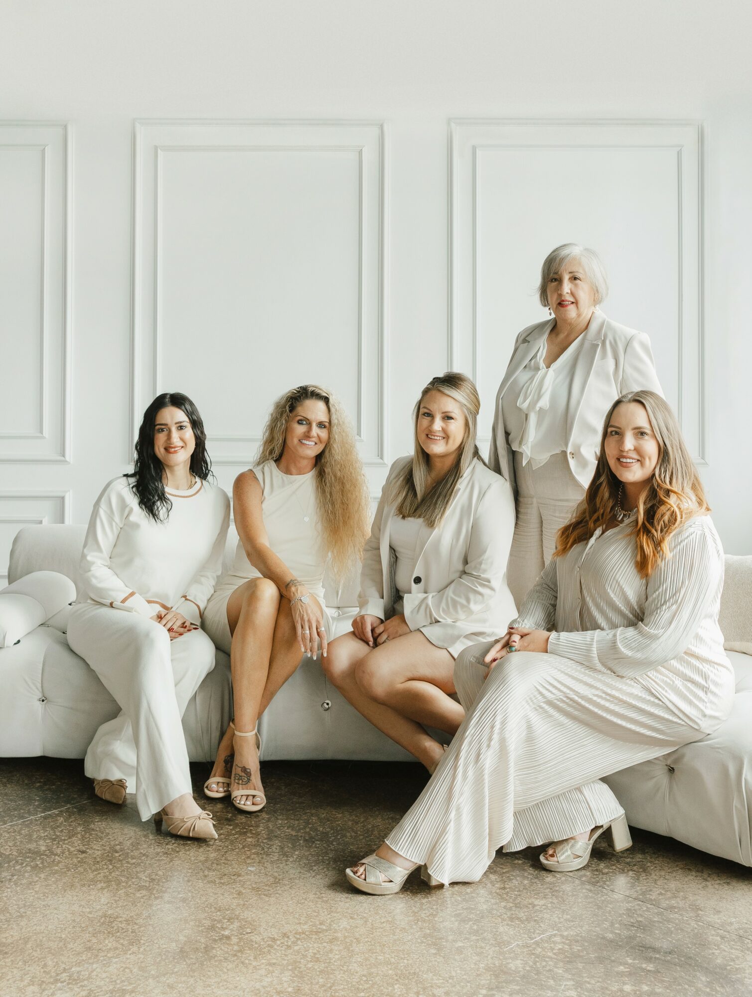 Five women sitting and standing on a white couch in a bright room with white walls, smiling, wearing white clothing.
