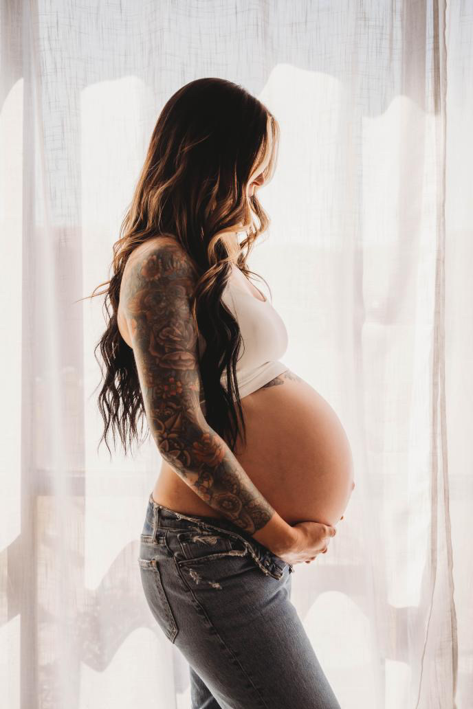 Pregnant woman with long hair, wearing a white top and jeans, standing sideways in front of curtains.