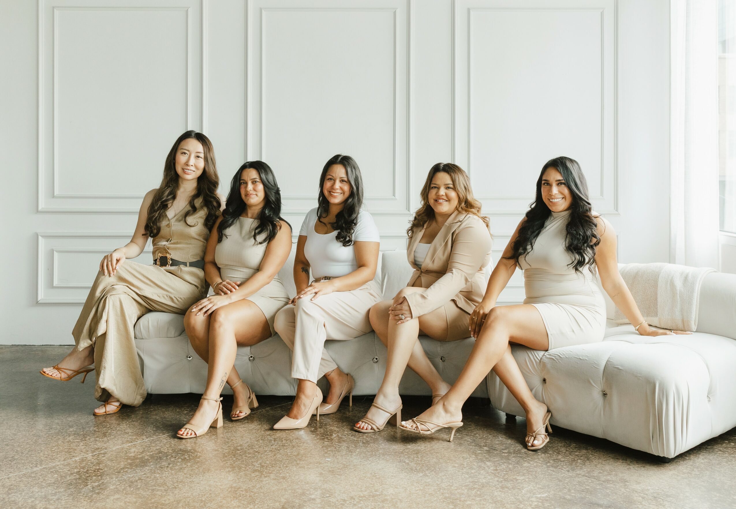 Five women sit on a white sofa in a bright, elegant room with white paneled walls and large windows.