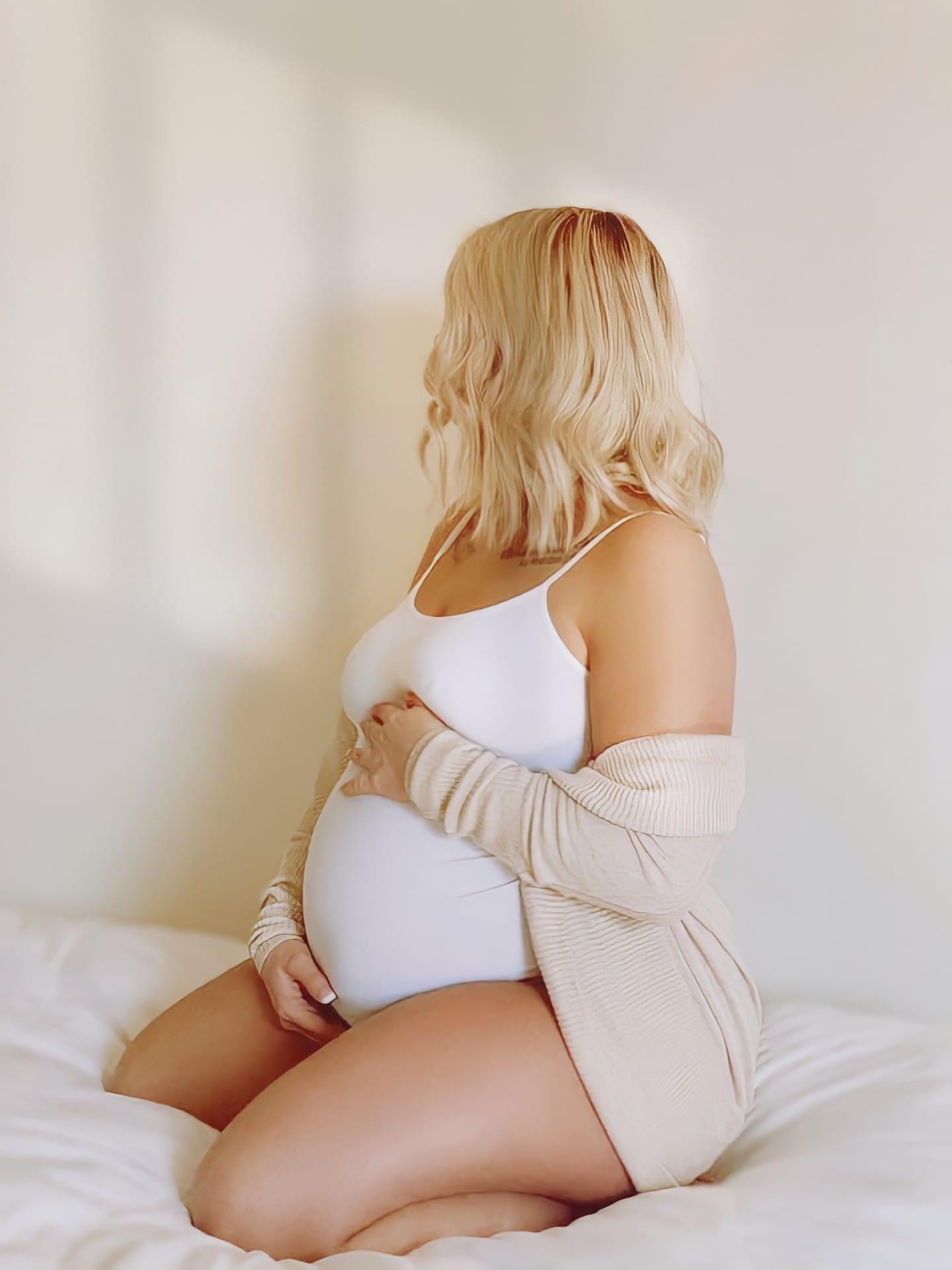 Pregnant woman with blonde hair sitting on a bed, wearing a white top and beige cardigan, facing sideways.