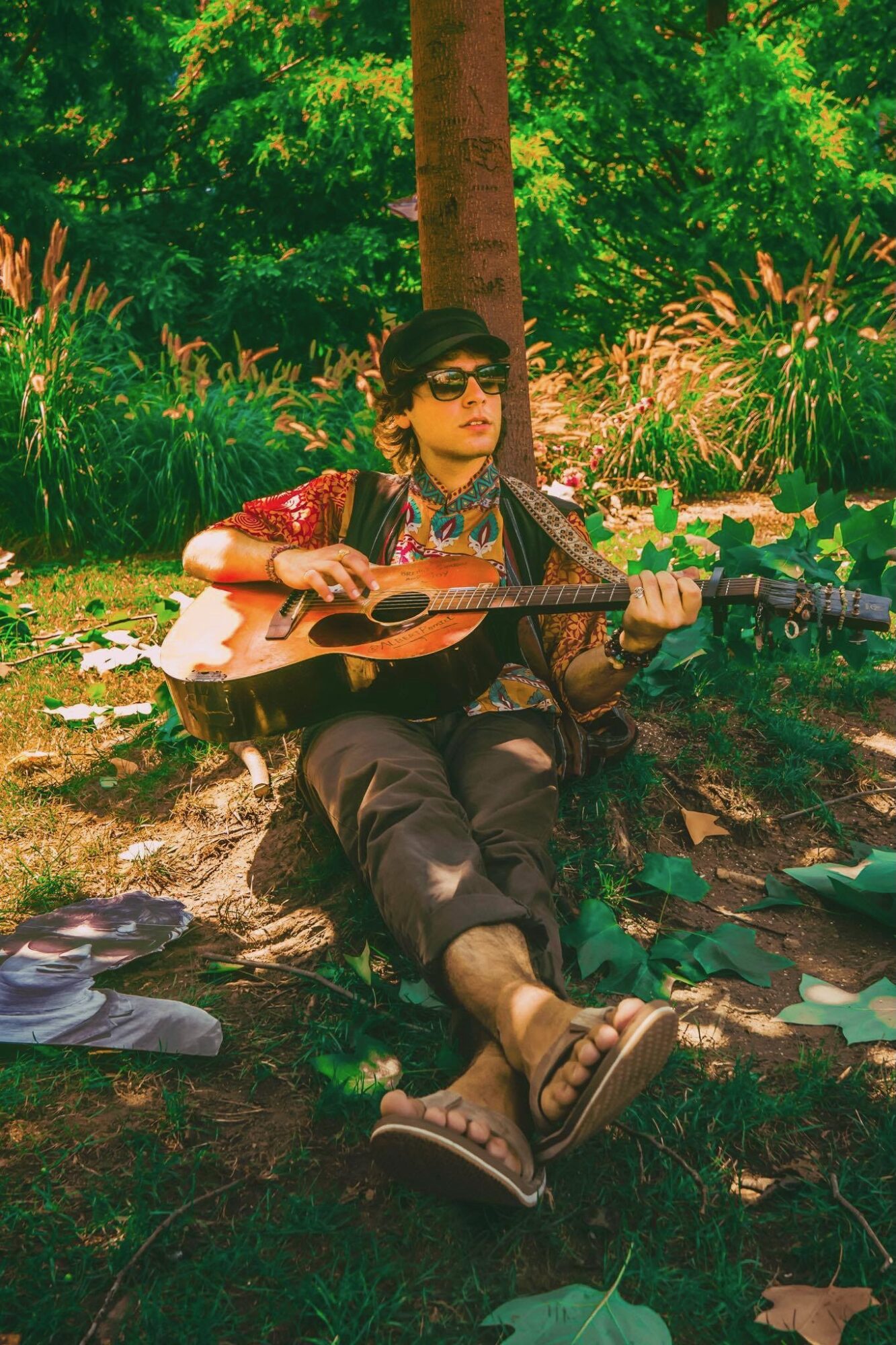 Person sitting on ground playing guitar outdoors with trees and plants in background.