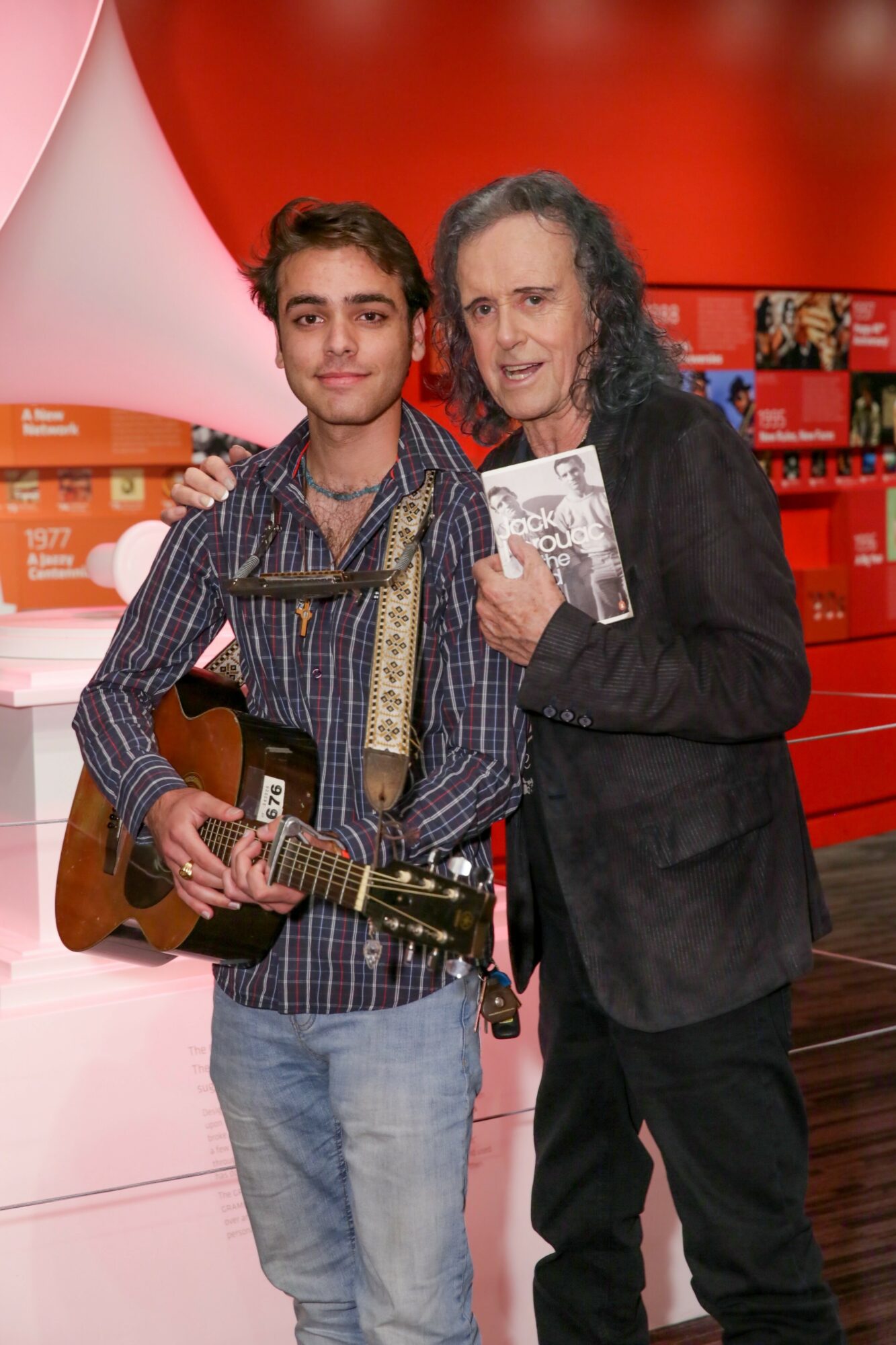 Young man with guitar and older man holding a magazine, standing together in a colorful indoor setting.