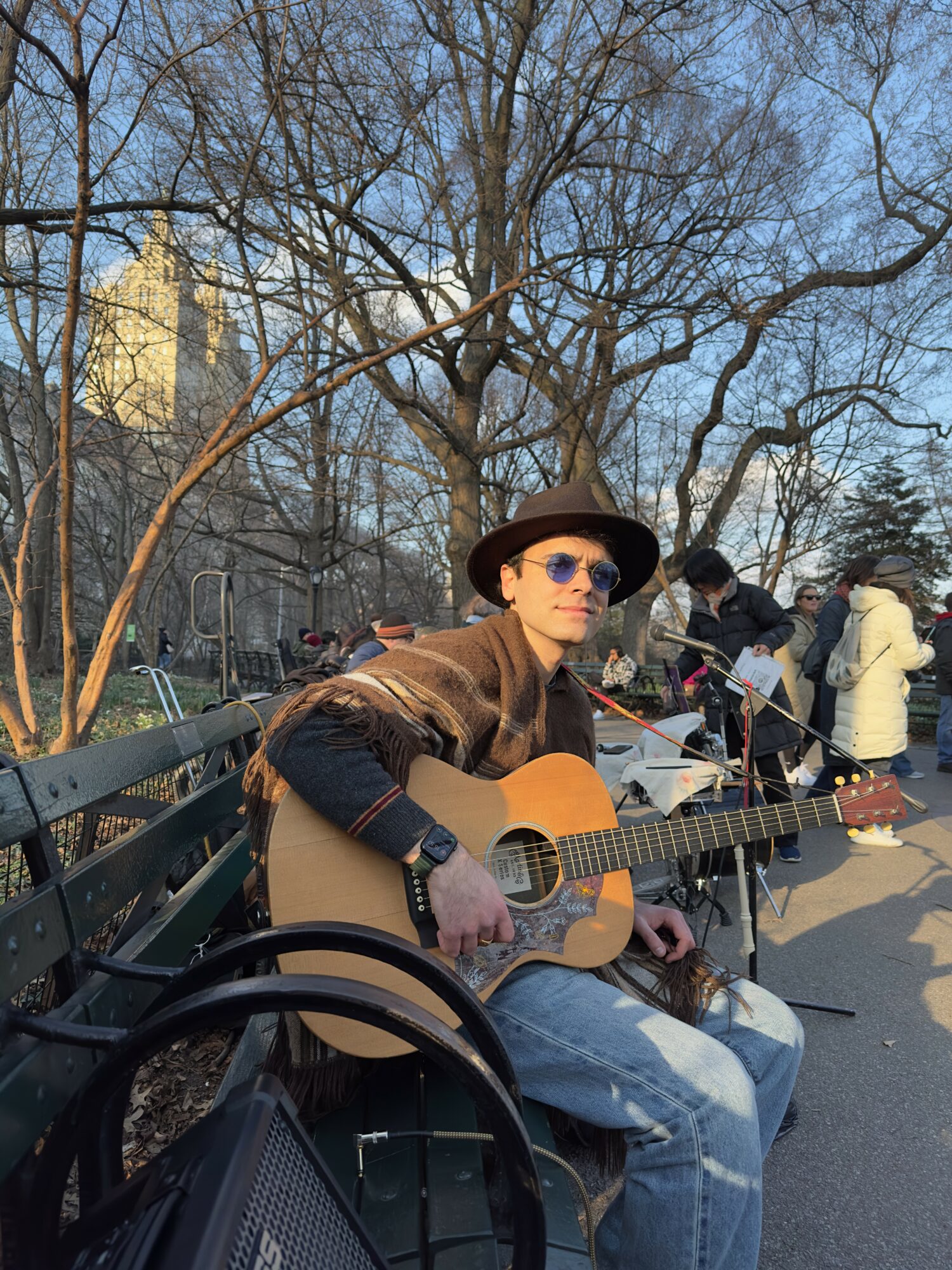 Man wearing sunglasses and a hat playing guitar on park bench with people in background, trees, and buildings.
