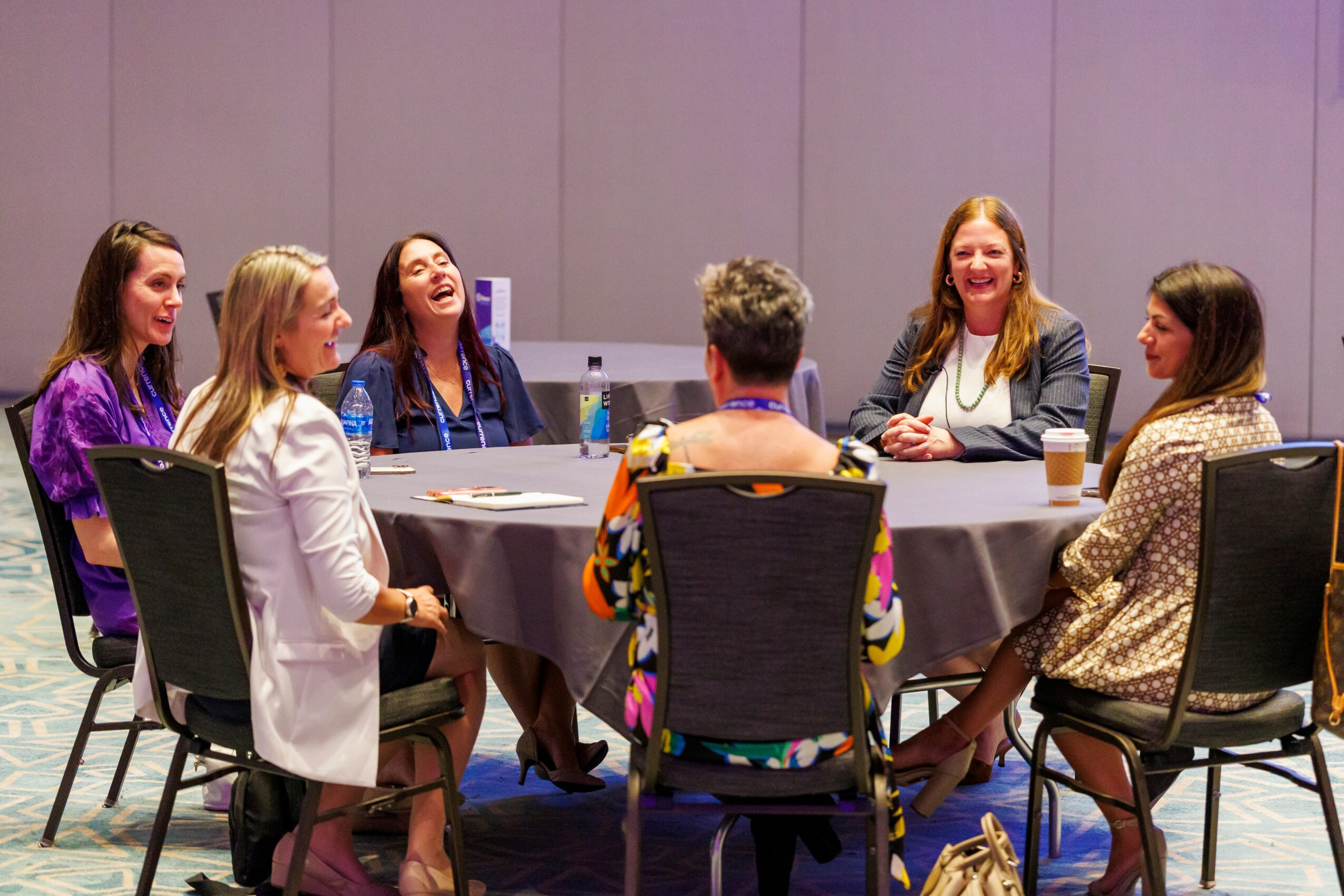 Seven women sit around a round table in a meeting room, engaged in conversation and smiling.